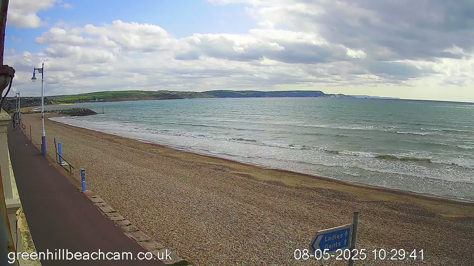 A pebble beach stretches toward a calm sea under a partly cloudy sky, with hills visible in the distance.