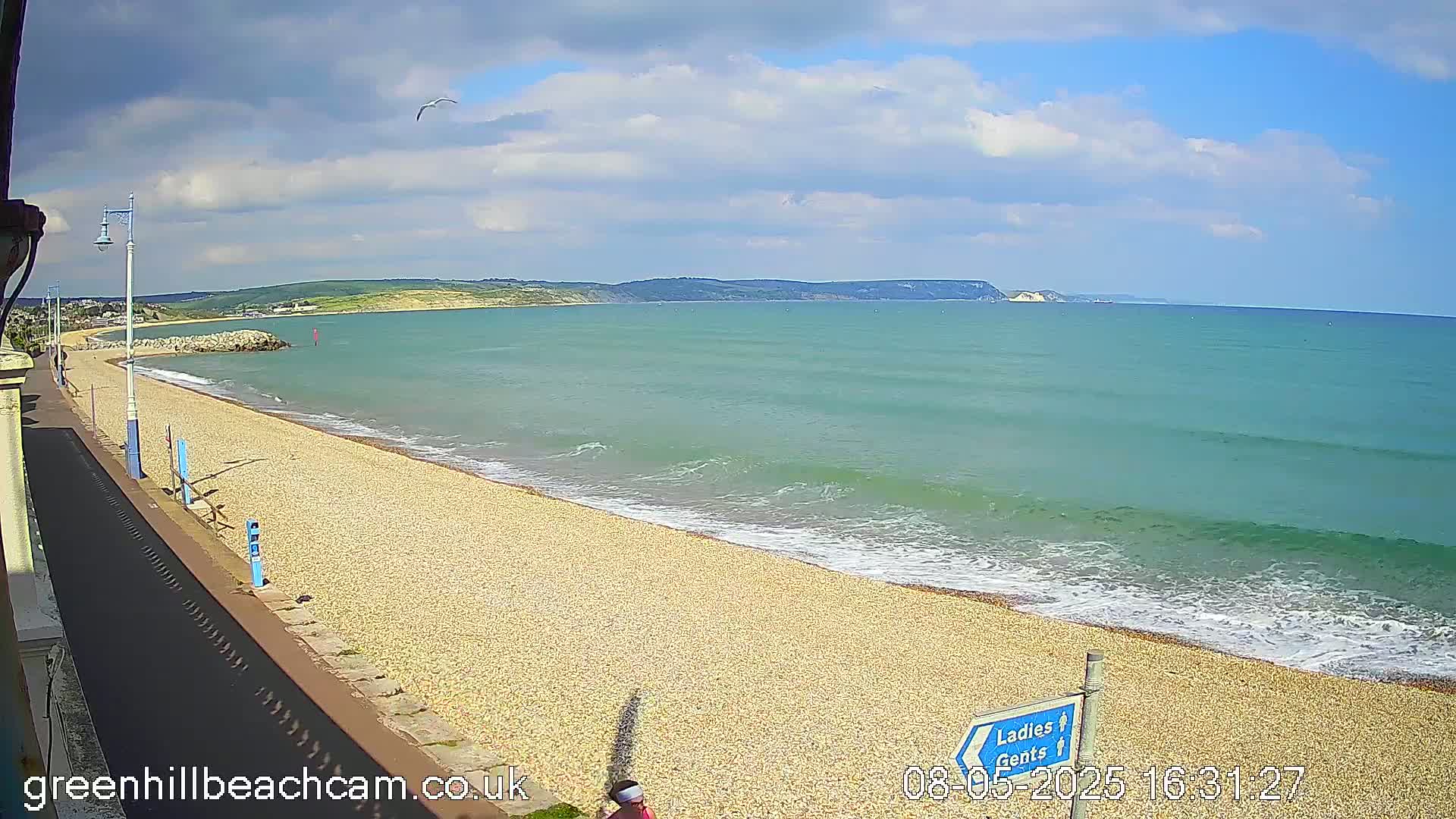 A pebble beach stretches along a calm, teal sea under a partly cloudy sky, with a distant headland visible.