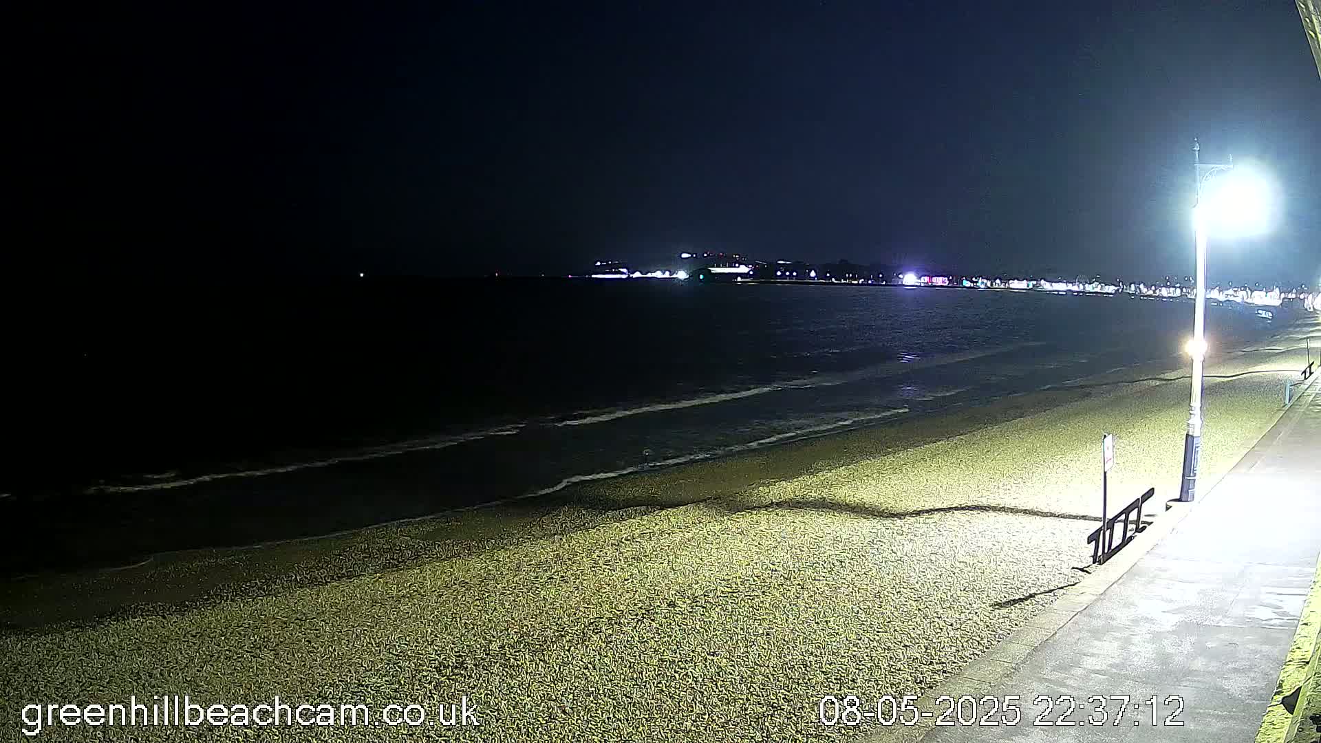 A dark, pebbled beach is illuminated by a streetlight at night, with gentle waves lapping the shore under a clear sky.