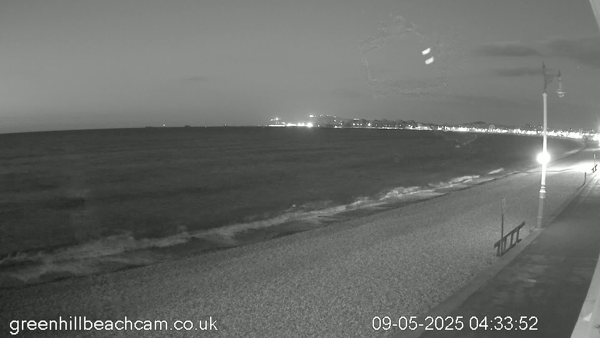 A dark, nighttime view of a pebble beach with gentle waves lapping the shore, and a town with lights visible in the distance.