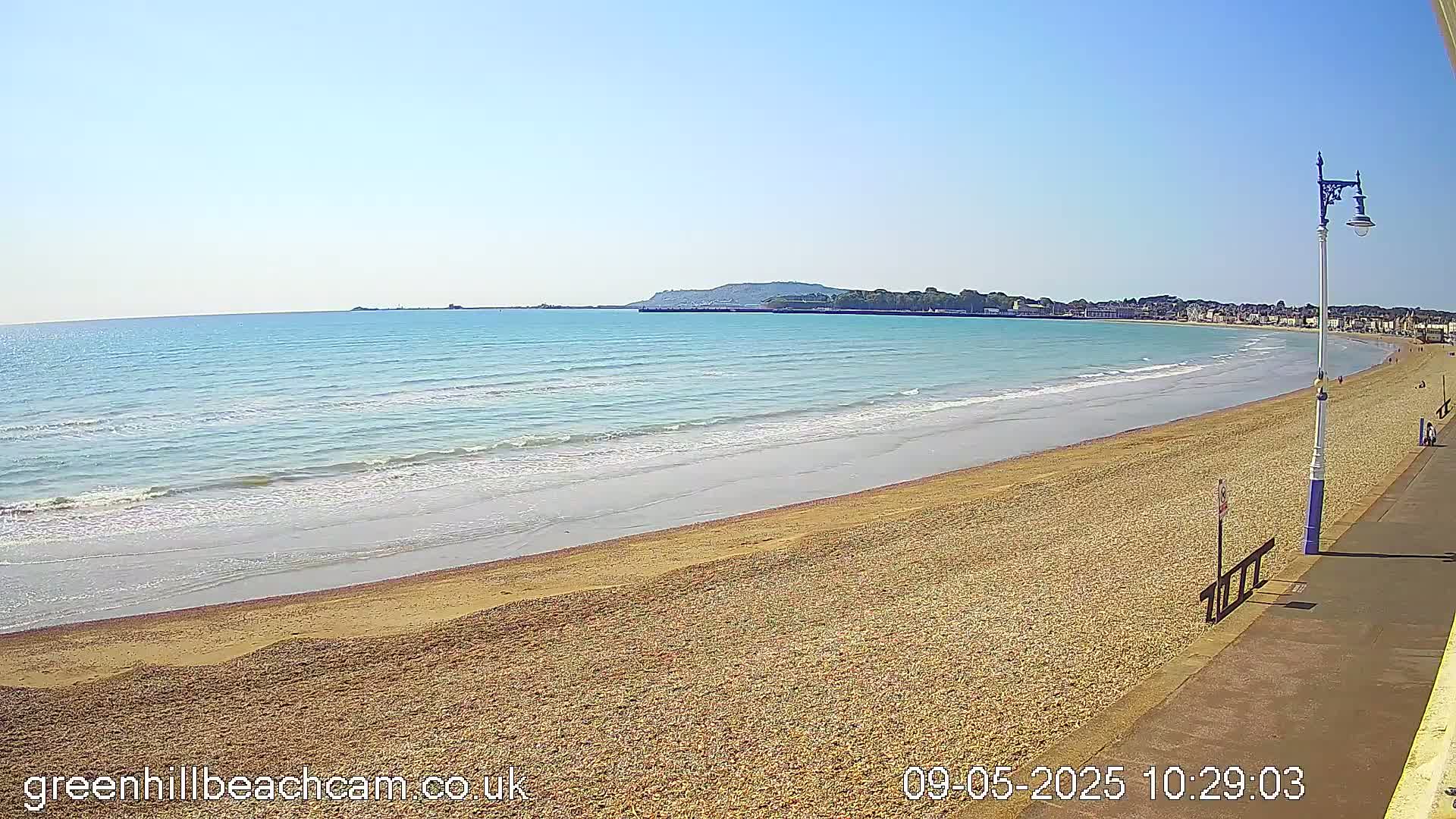 A sandy beach with gentle waves stretches along a calm, blue ocean under a clear, sunny sky, next to a paved walkway and distant town.