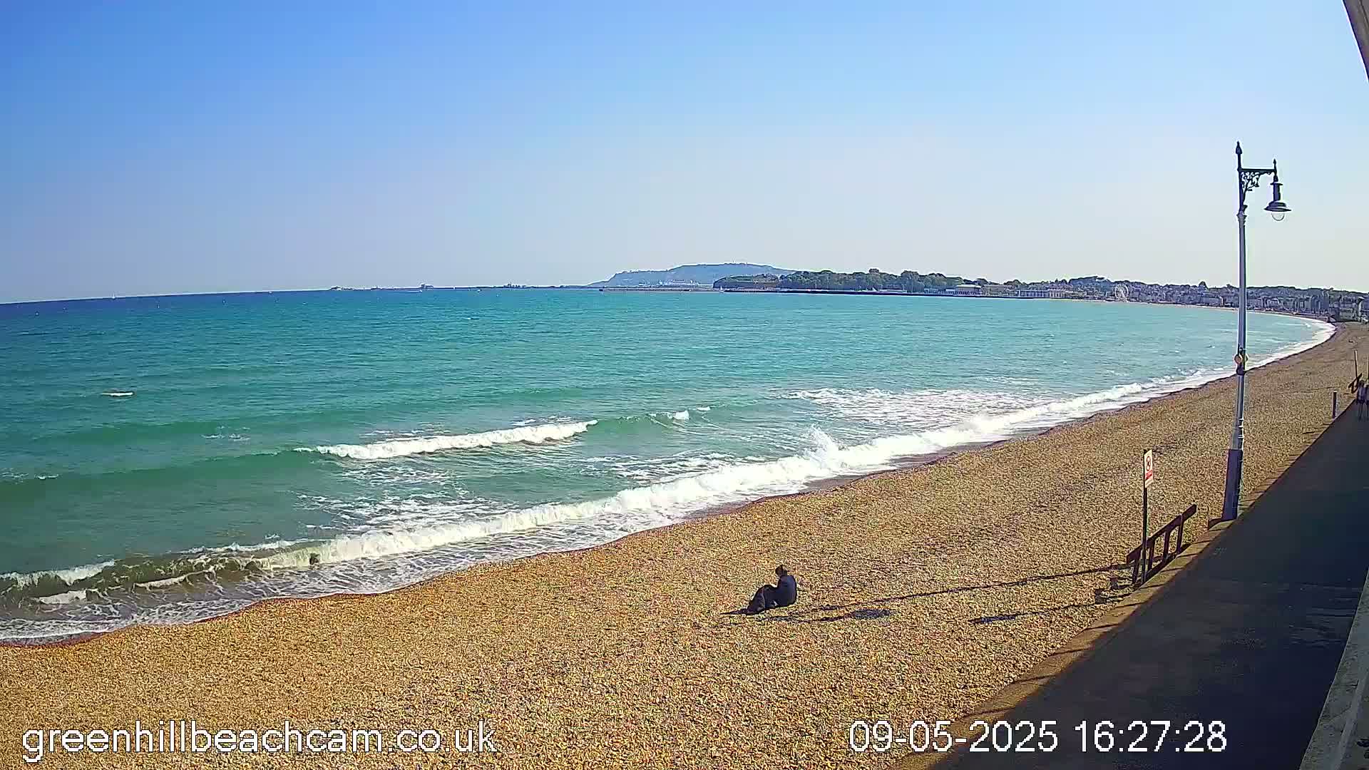A person sits on a pebble beach next to a calm, turquoise ocean under a clear blue sky.