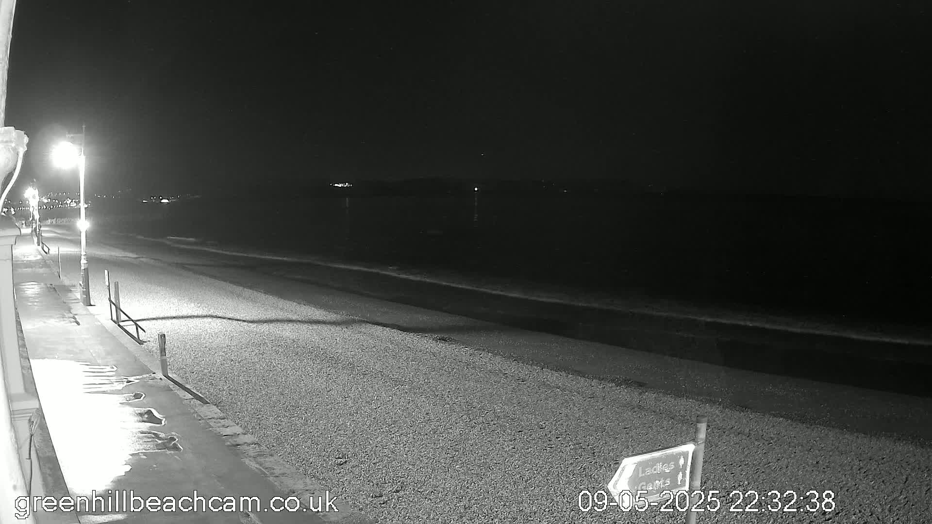 A dark, nighttime beach scene shows a pebble shore with gentle waves lapping at the edge, under a clear, starless sky, illuminated by streetlights along a paved walkway.
