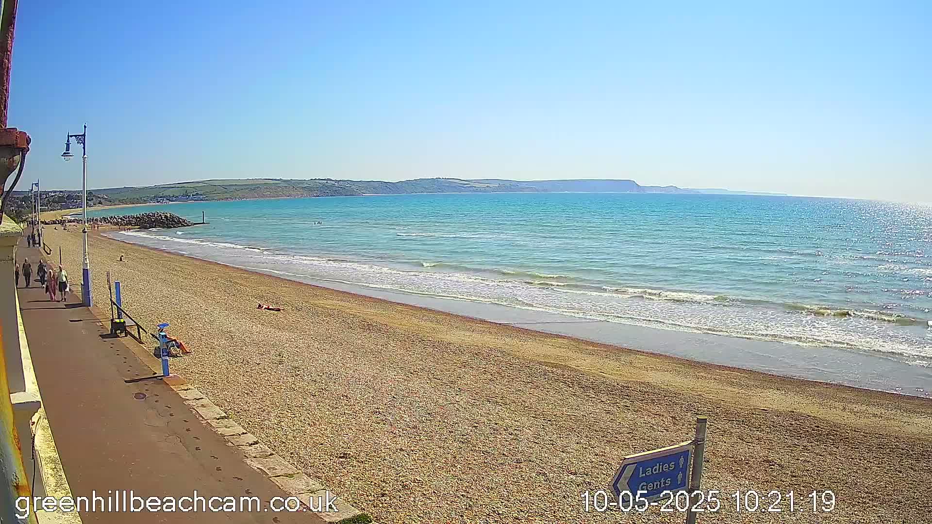 A sunny day at a pebble beach shows calm ocean waves lapping the shore, with people walking along a paved path and relaxing on the sand.