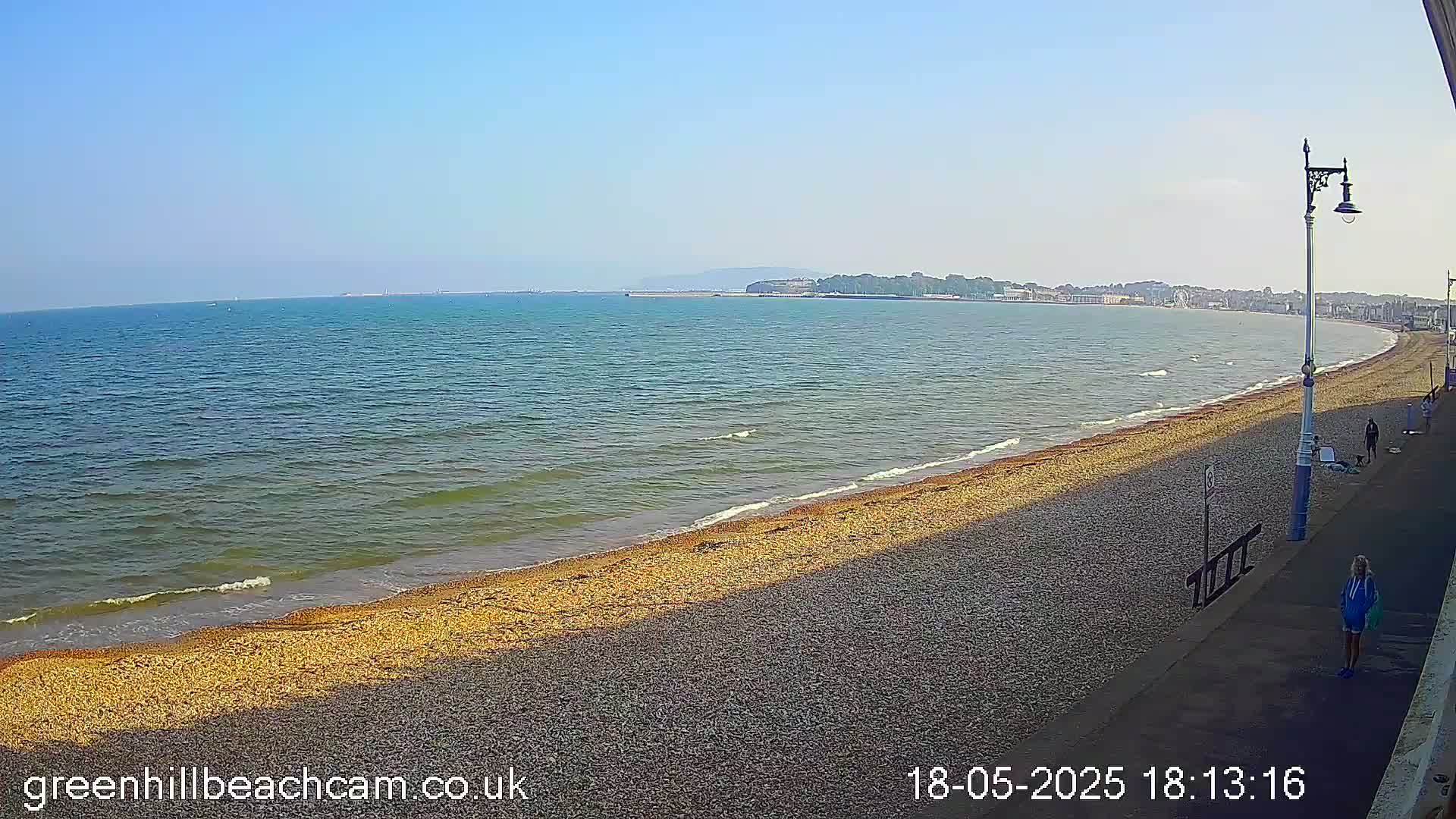 A pebble beach stretches along a calm, blue sea under a clear, sunny sky, with a few people walking along a promenade.