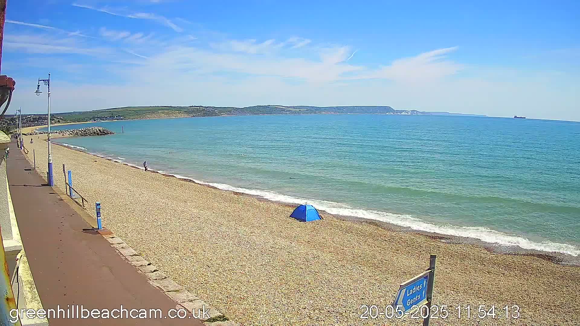 A pebble beach with a small blue tent and a few people, under a sunny sky, is situated next to a calm, turquoise sea and backed by a hilly coastline.