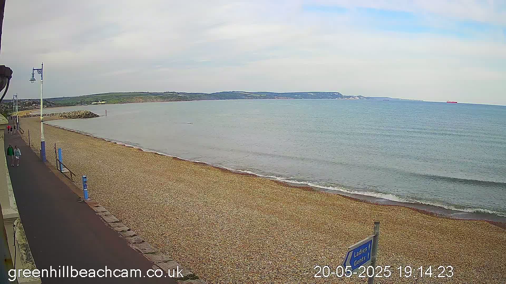 A pebble beach with a paved walkway and people walking, extending to a calm sea under a cloudy sky, with distant hills and a few ships visible on the horizon.