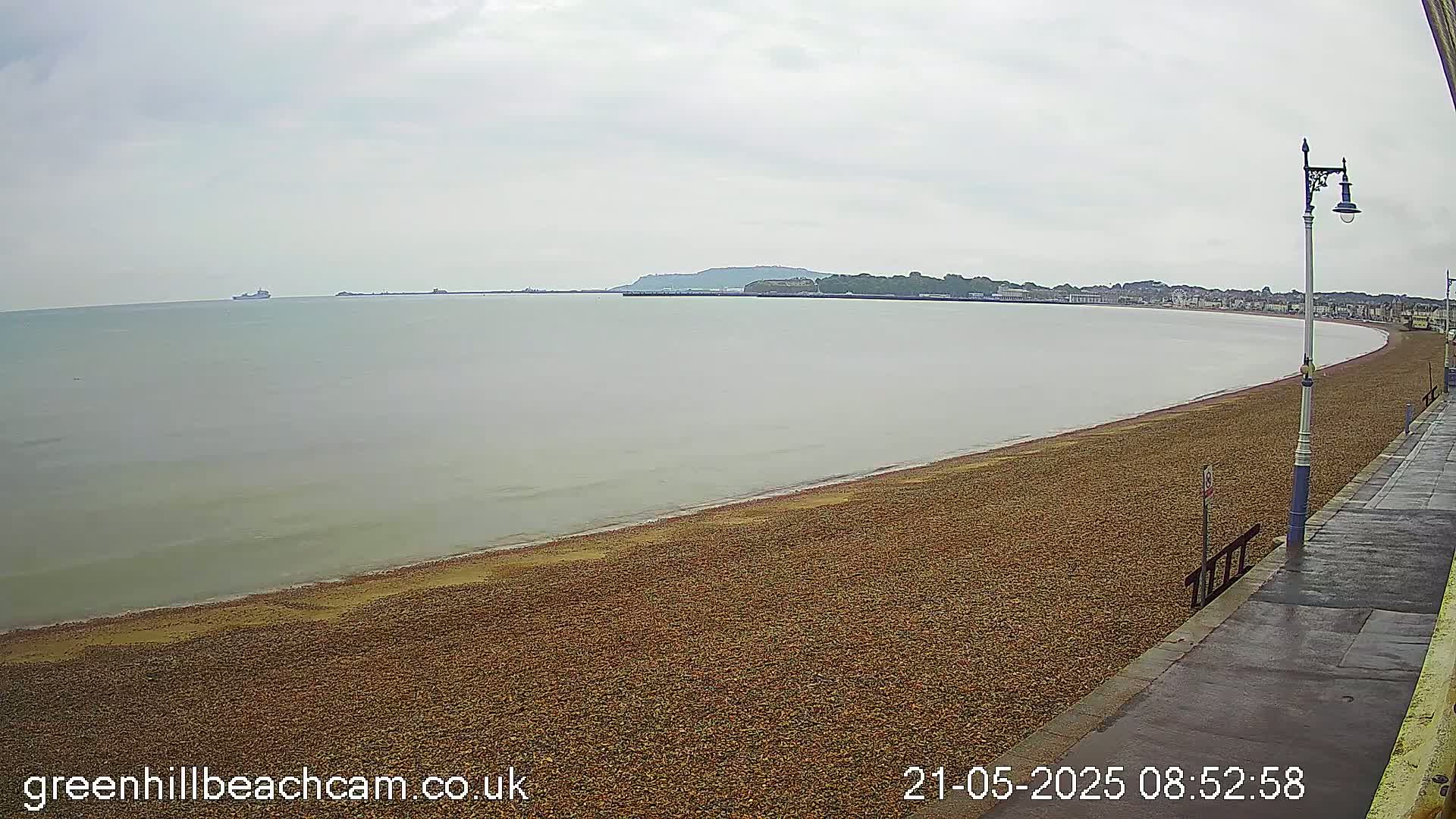 A pebble beach curves along a calm sea under an overcast sky, with a distant town visible across the water.