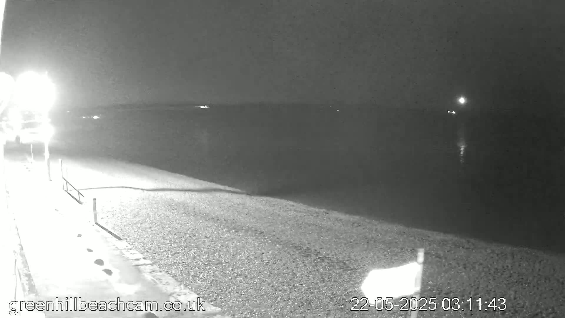 A nighttime view shows a dark, calm body of water next to a pebbled shoreline, lit by a single bright lamp.