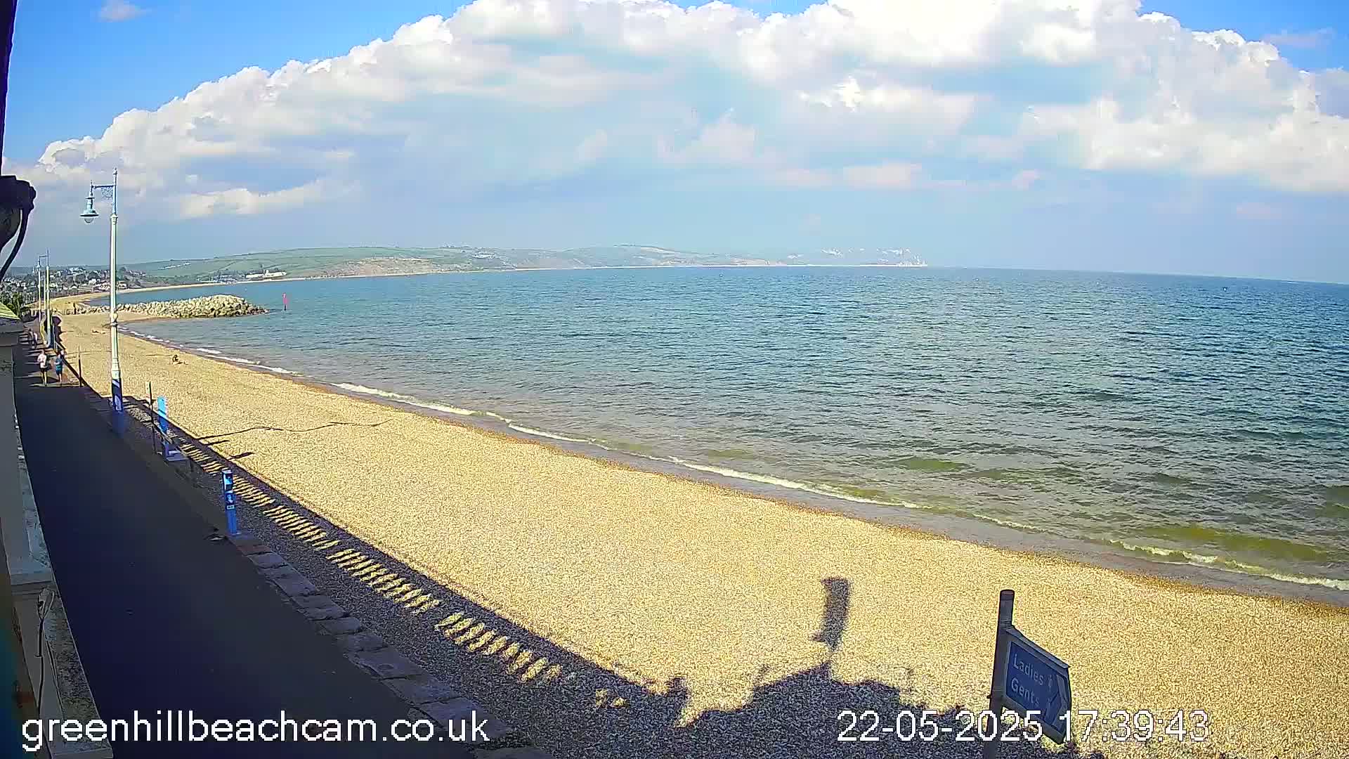 A partly cloudy sunny day shows a pebble beach gently sloping to a calm sea, with a paved walkway and distant hills visible.