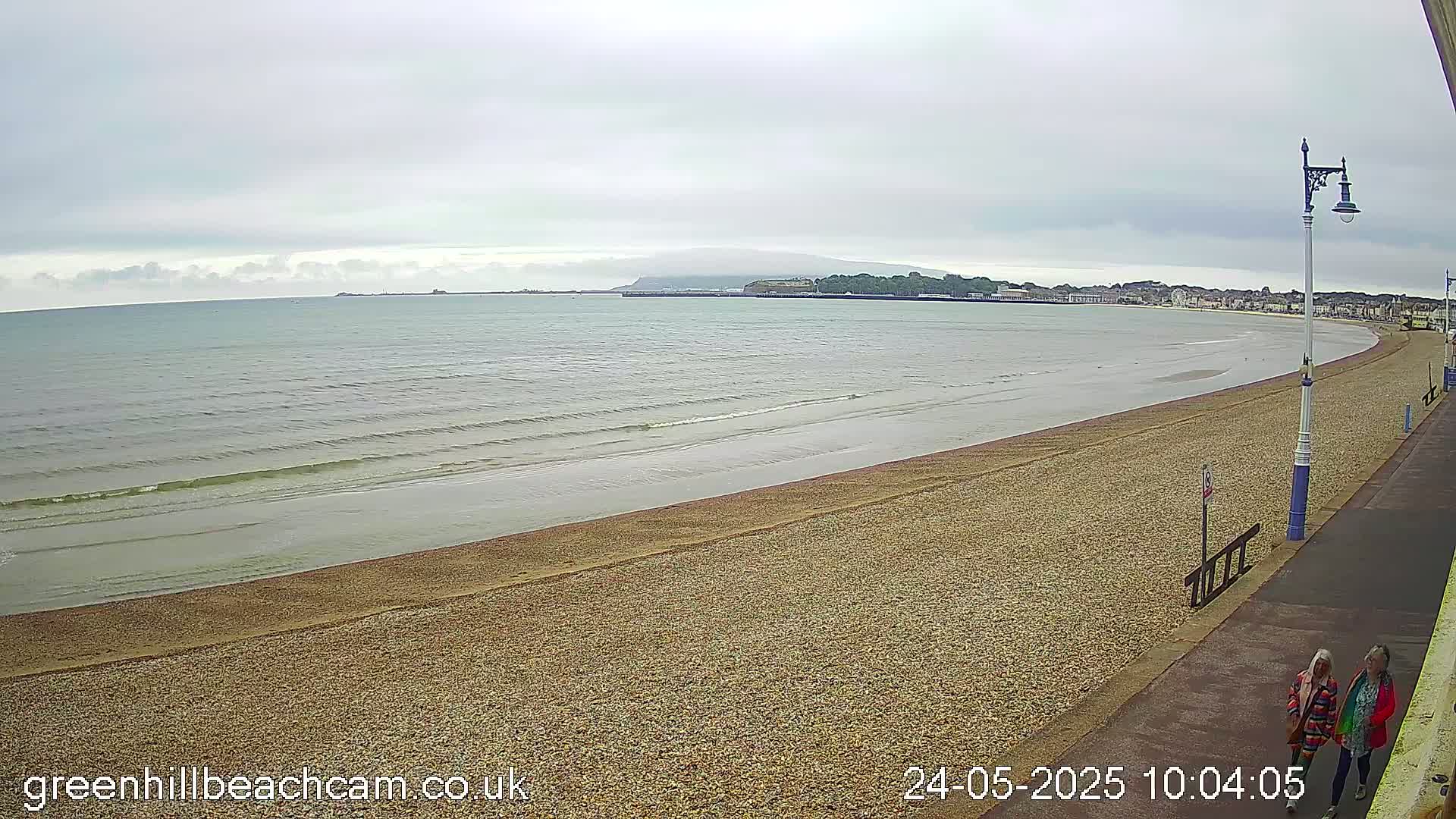 A long, sandy beach stretches to a calm sea under an overcast sky, with a few people walking along a paved path next to it.