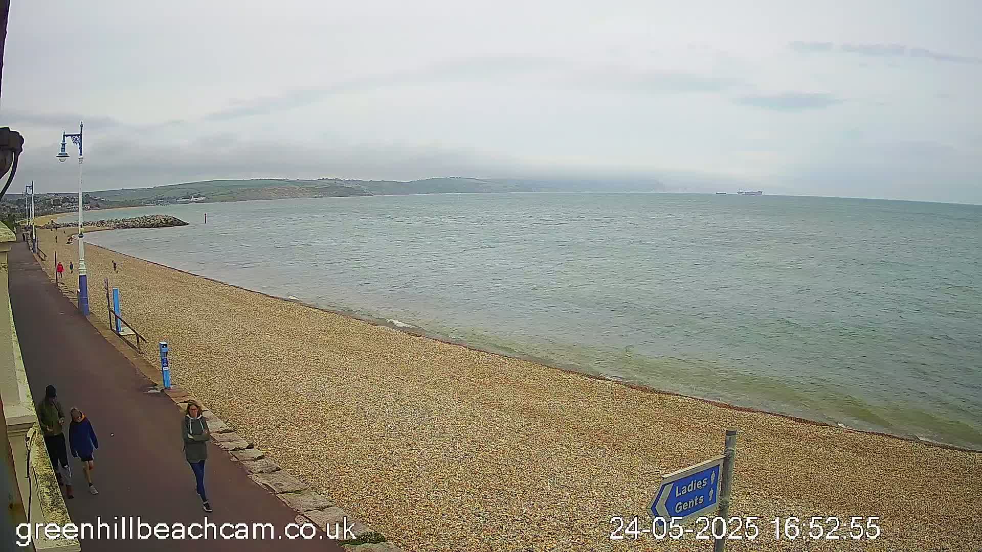 A pebble beach stretches along a calm sea under an overcast sky, with a few people walking along a paved path beside it.