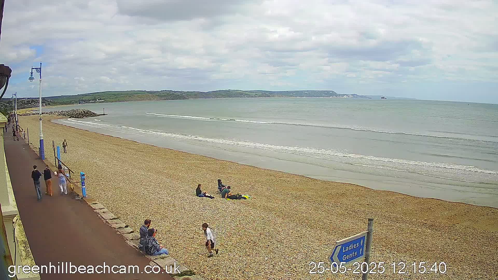A pebble beach with a few people walking and sitting, under a partly cloudy sky, with gentle waves lapping the shore.
