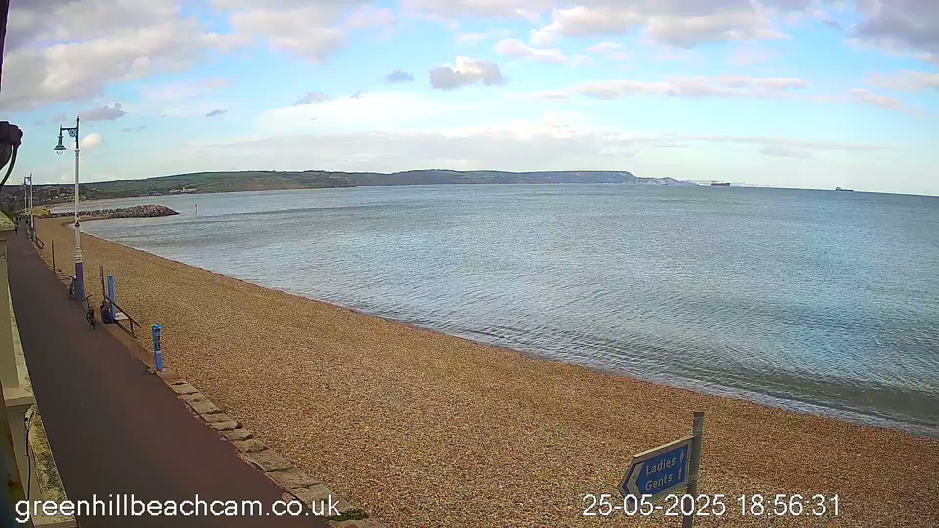 A pebble beach stretches along a calm sea under a partly cloudy sky, with a paved walkway and distant hills visible.
