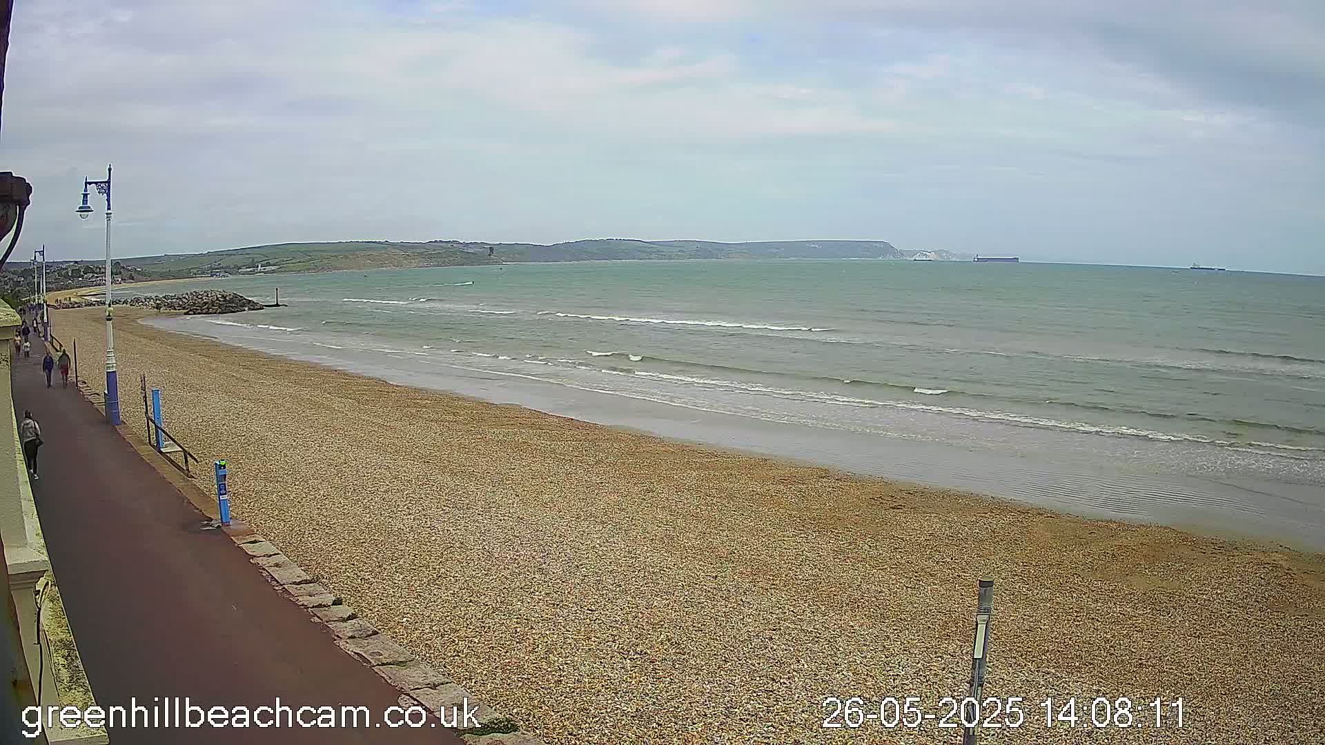 A pebble beach with a paved walkway and several people walking along it stretches toward a calm sea under a cloudy sky.