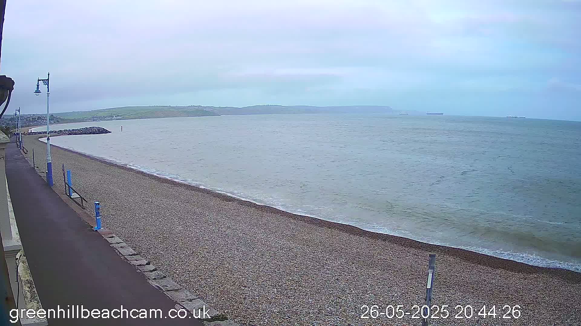 A pebble beach stretches along a calm sea under an overcast sky, with a paved walkway and lampposts visible along the shore.