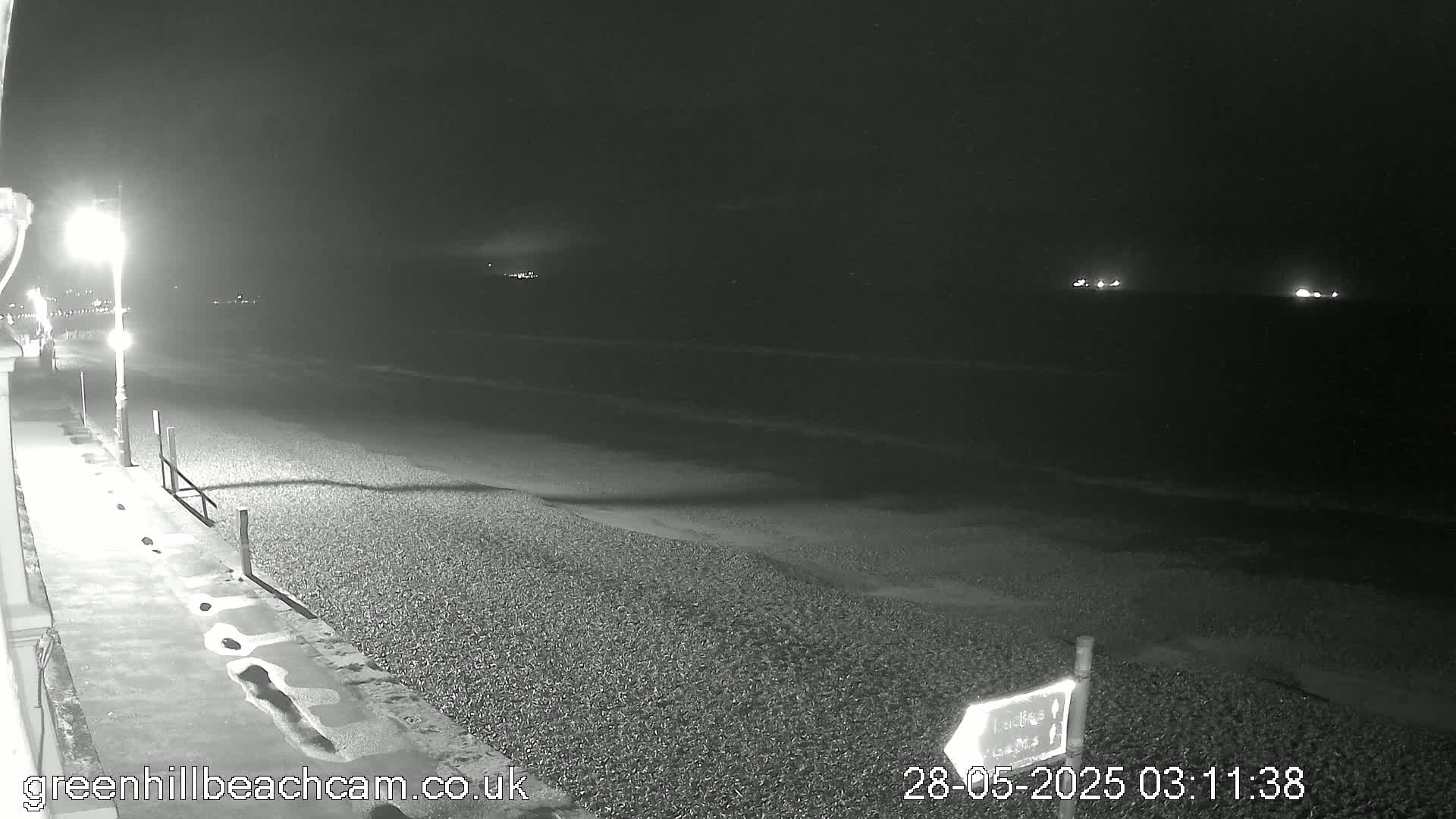 A nighttime view of a pebble beach, illuminated by a single streetlight, with calm waves and distant lights visible across the water.