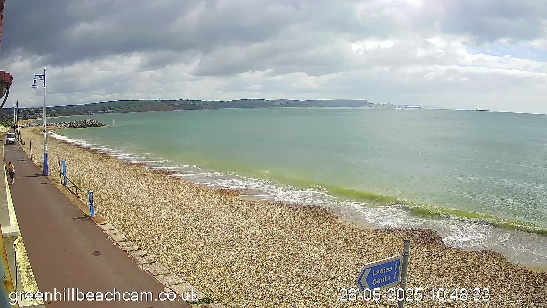 A pebble beach with gentle waves under a partly cloudy sky, a paved walkway along the beach, and a few people walking.