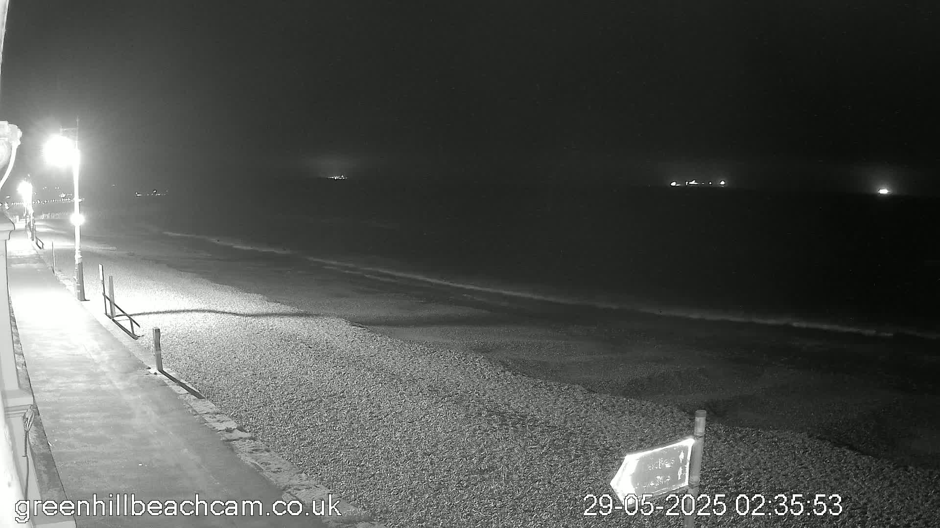 A dark, nighttime beach scene shows a pebble shore under a clear sky, with gently lapping waves and lit walkways nearby.