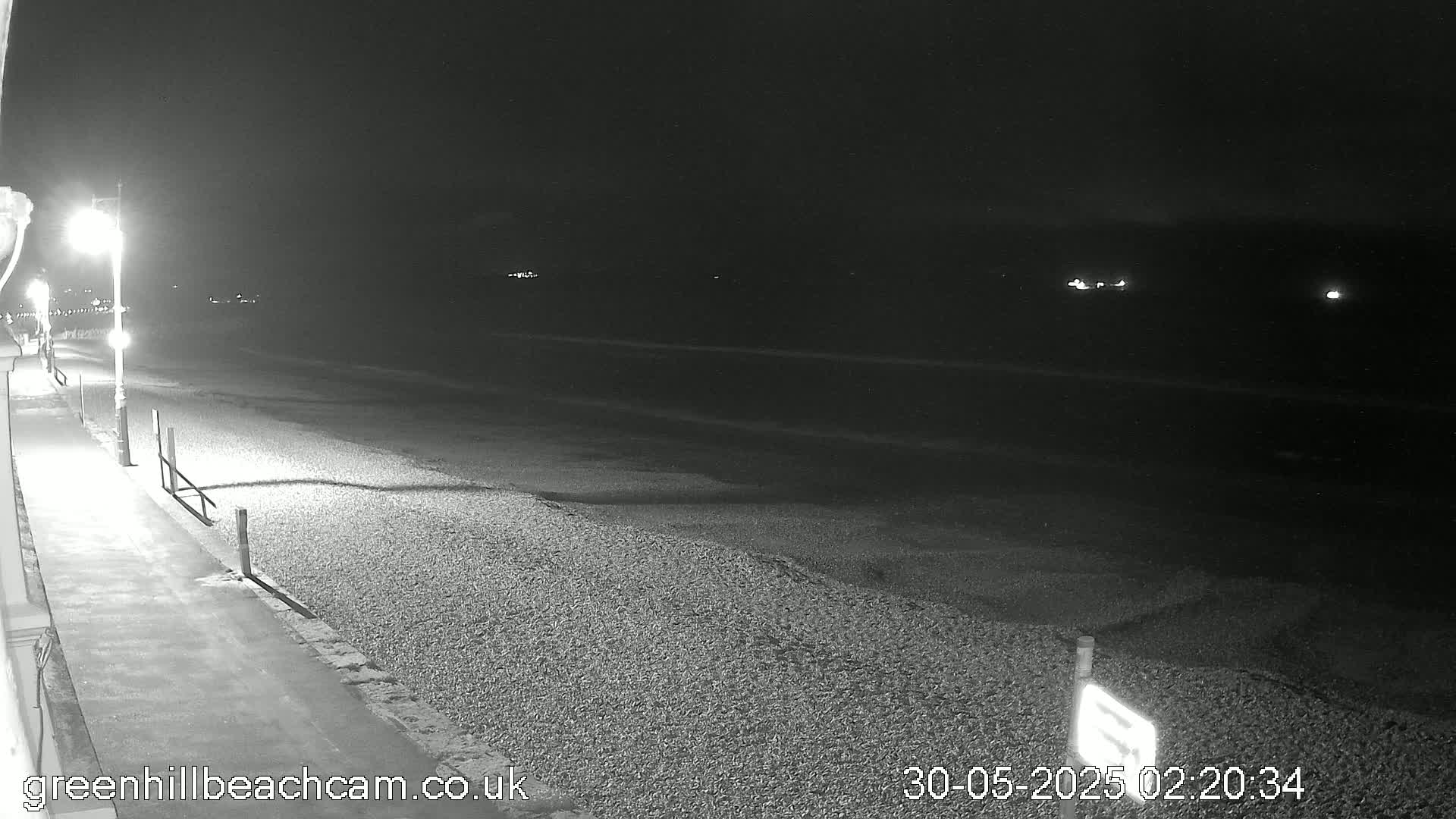 A nighttime view of a dark, pebble beach next to a paved walkway lit by streetlights, with a calm sea in the background under a clear sky.