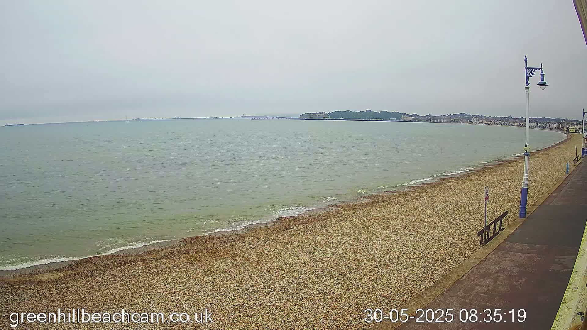 A pebble beach stretches along a calm, gray sea under an overcast sky, with a town visible in the distance.