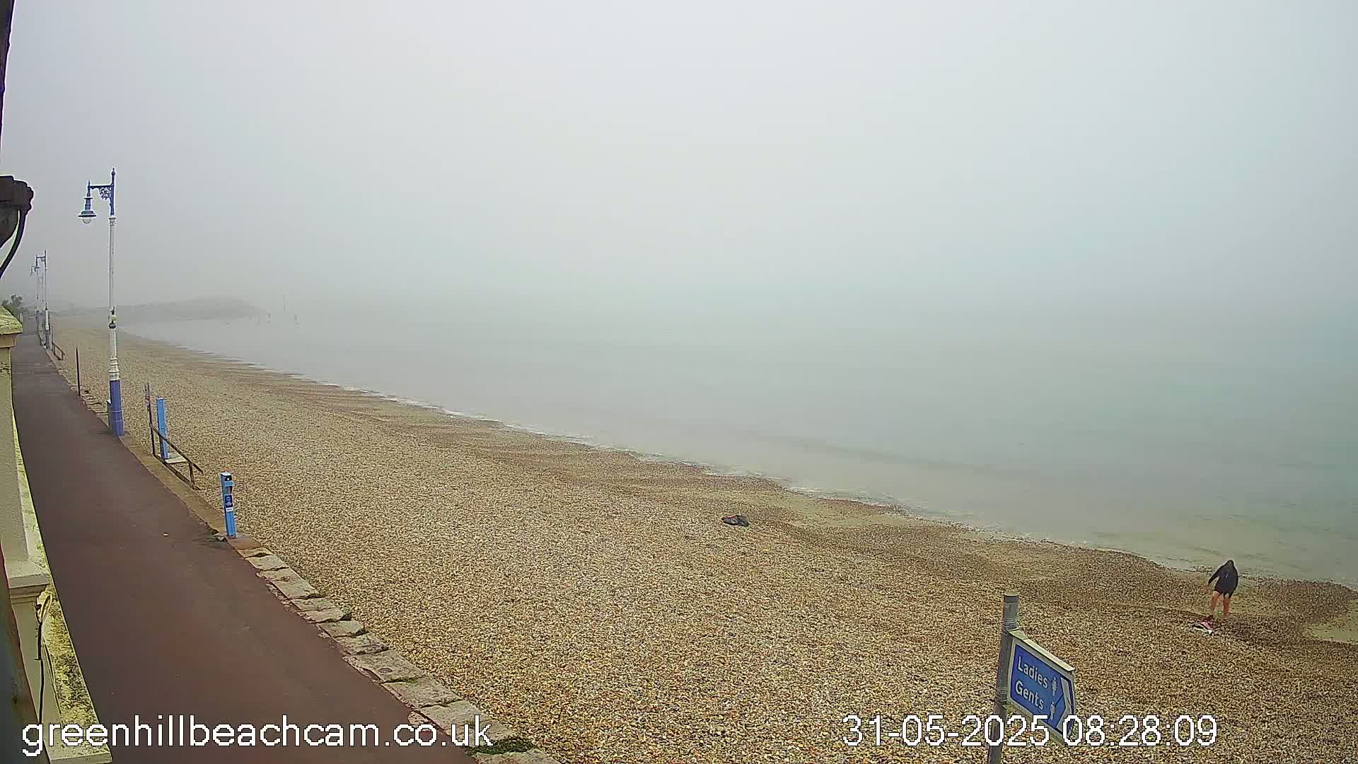A person walks on a pebble beach next to a calm, foggy sea, with a paved walkway and lampposts lining the shore.