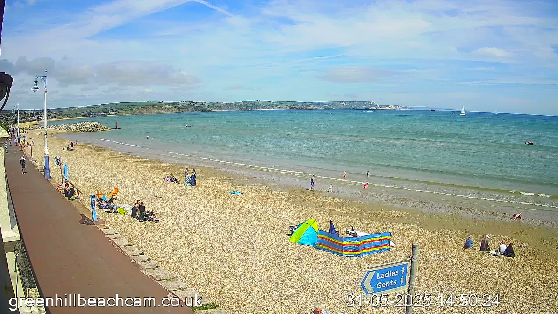 A sunny day at a pebble beach shows people relaxing and swimming in calm, clear water with a distant coastline visible under a partly cloudy sky.