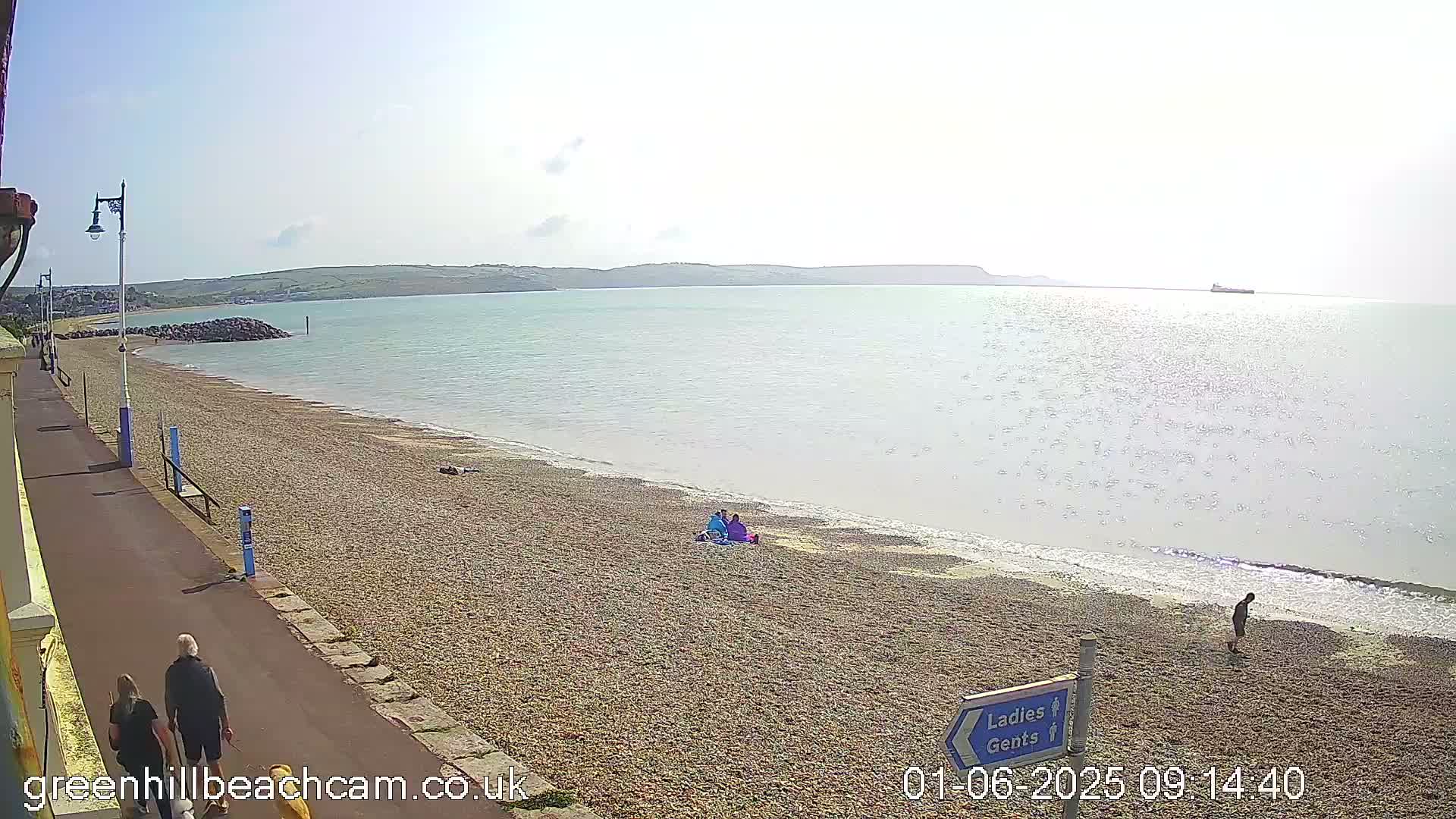 A pebble beach stretches along a calm sea under a bright, sunny sky, with several people walking and sitting on the shore.