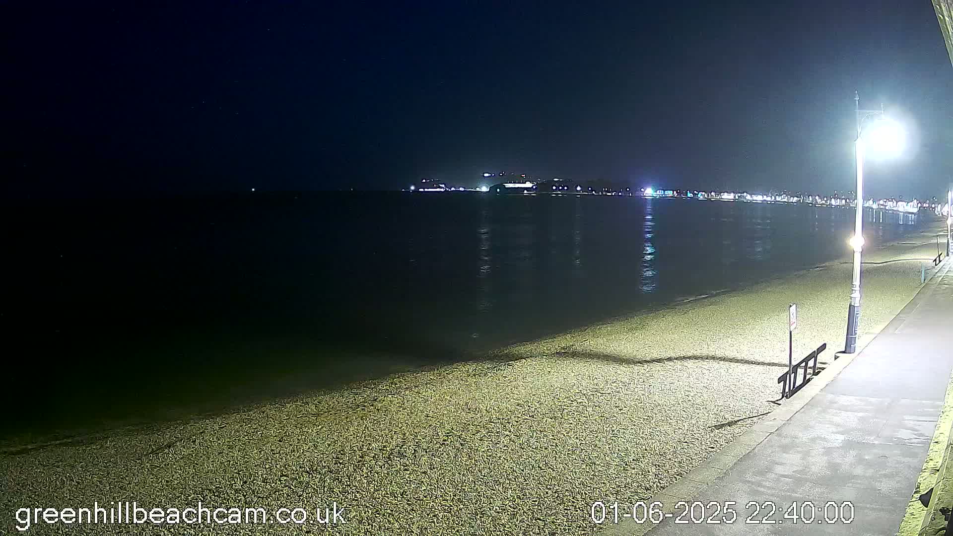 A dark, nighttime beach scene shows a calm, dark body of water reflecting distant lights, a pebbled shoreline, and a paved walkway with a lamppost.