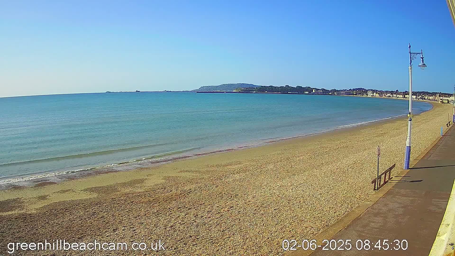 A sandy beach stretches along a calm, blue sea under a clear sky, with a promenade and distant town visible.