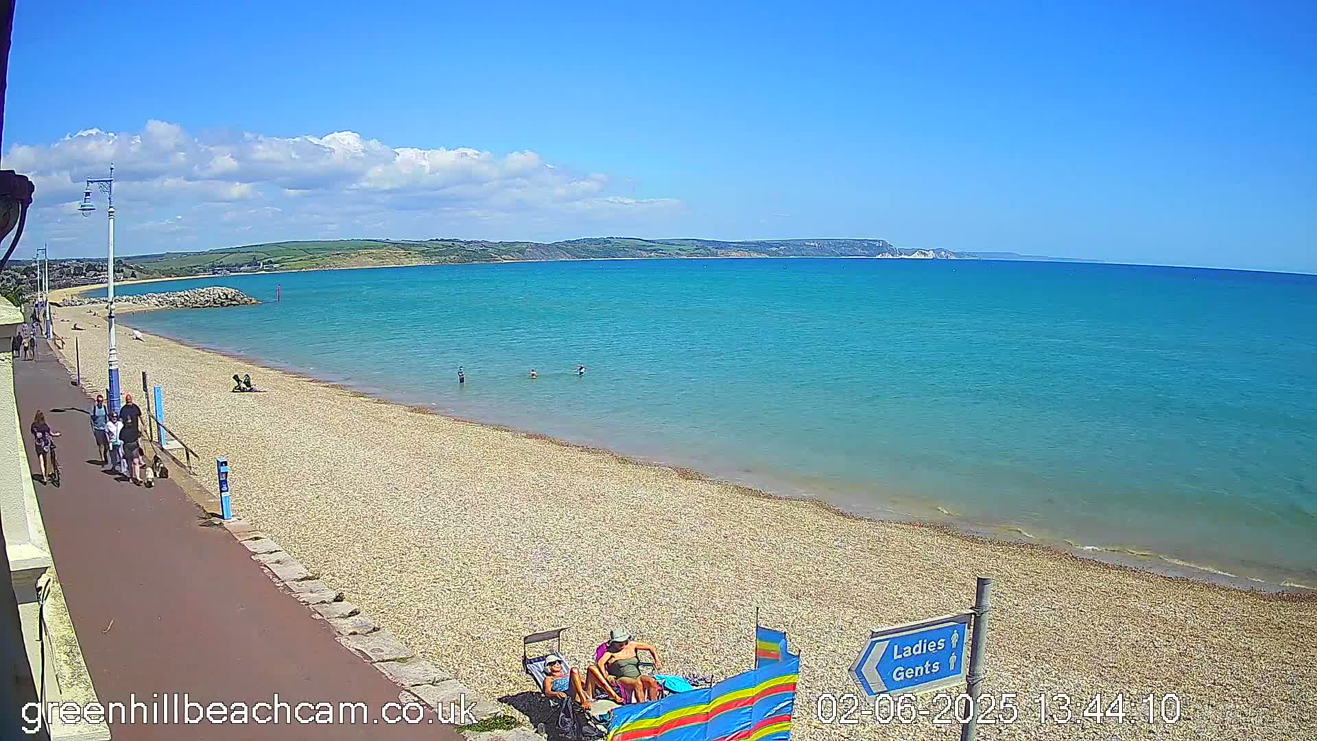 A sunny day at a pebble beach shows people walking along a paved path, some swimming in the clear turquoise water, and others relaxing on the shore.