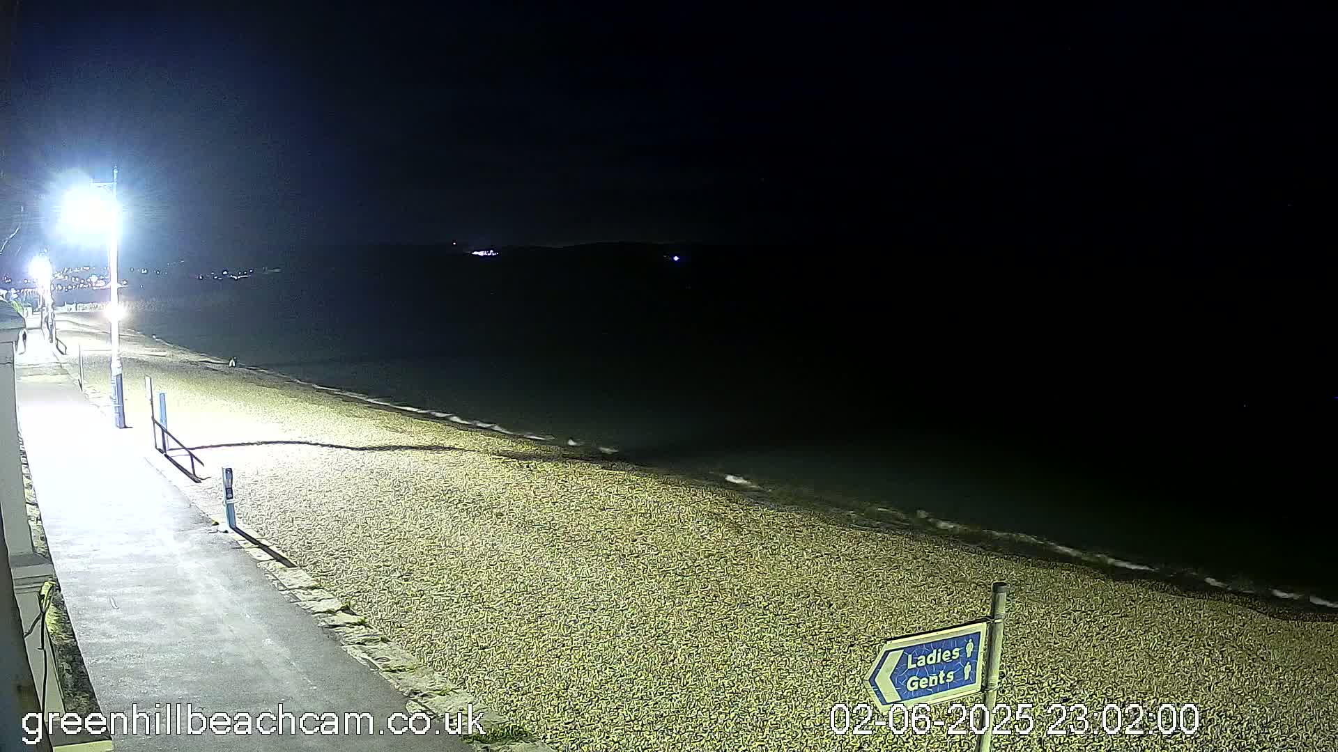 A dark, nighttime view of a pebble beach with a paved walkway and lampposts, gently lapping waves visible at the water's edge.