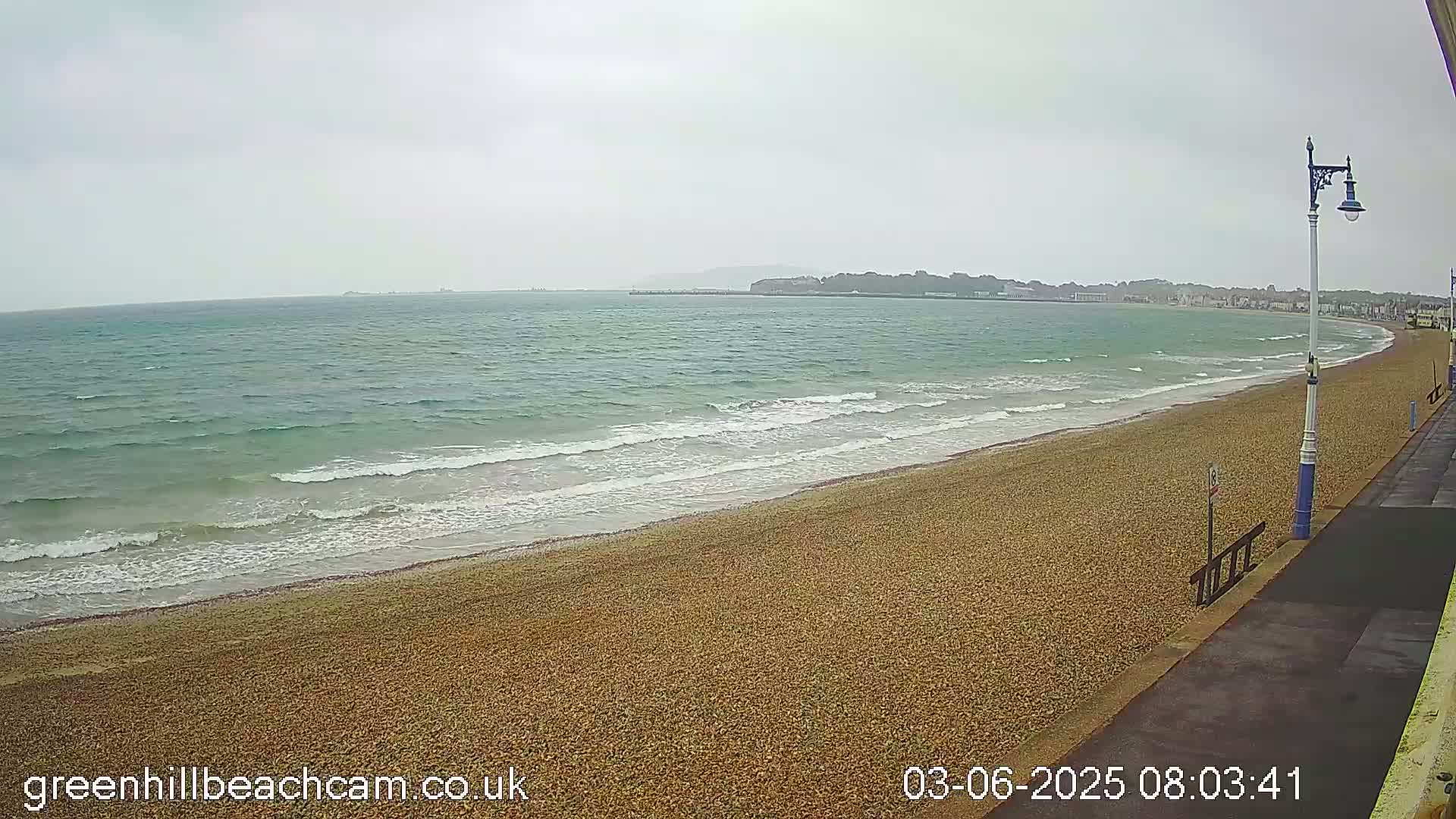 A pebble beach with gentle waves on a cloudy day stretches towards a distant shoreline.