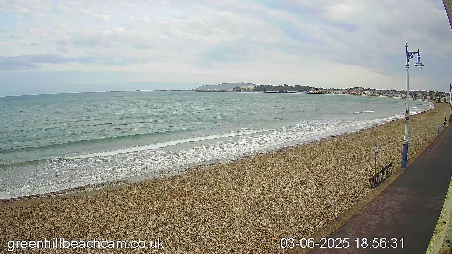 A pebble beach with gentle waves under a cloudy sky stretches alongside a paved walkway toward a town in the distance.