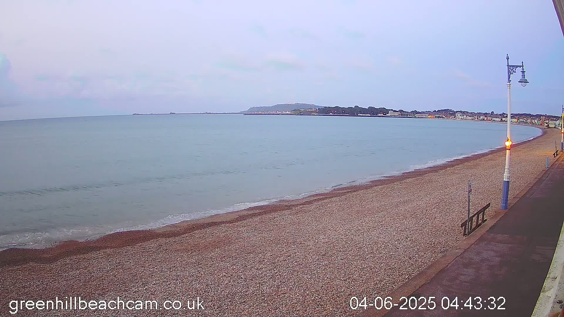 A calm, overcast morning reveals a pebble beach curving along a shoreline with a town visible in the distance.