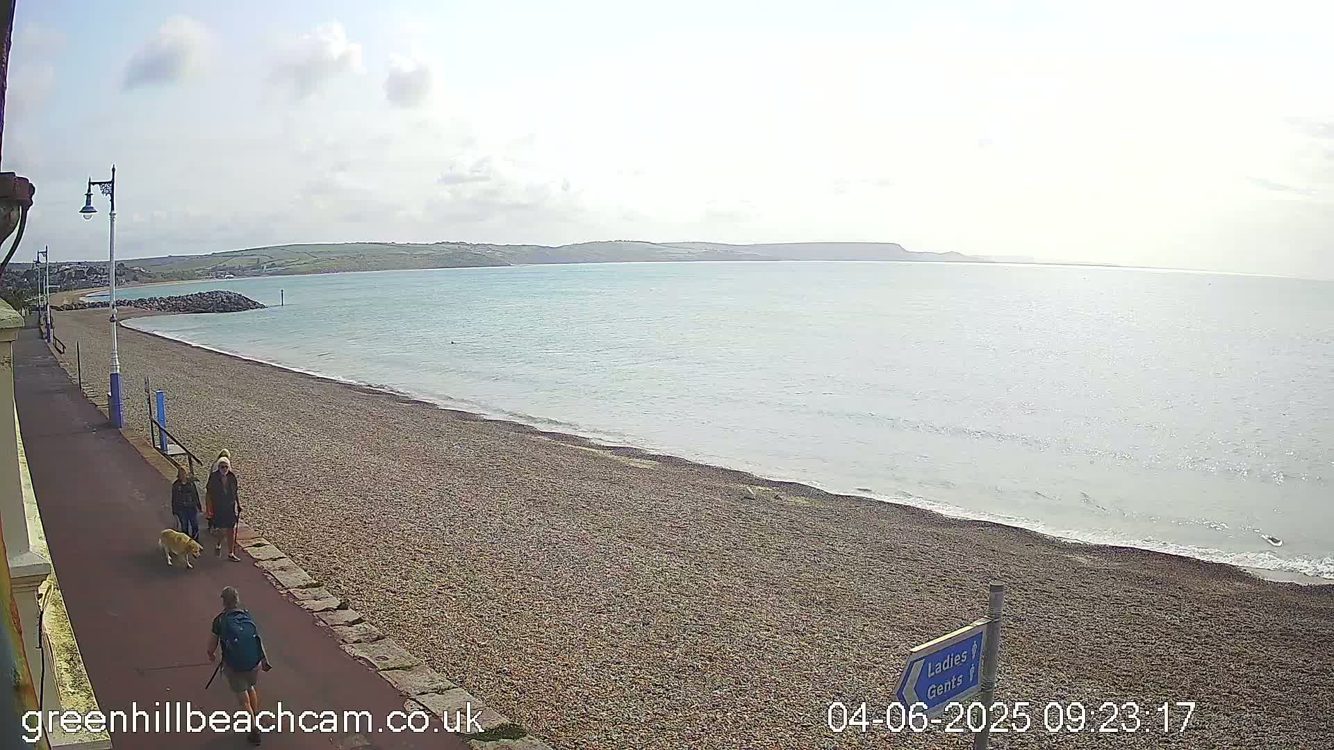 A pebble beach with a calm sea under a partly cloudy sky, features a paved walkway with people and a dog walking along it.