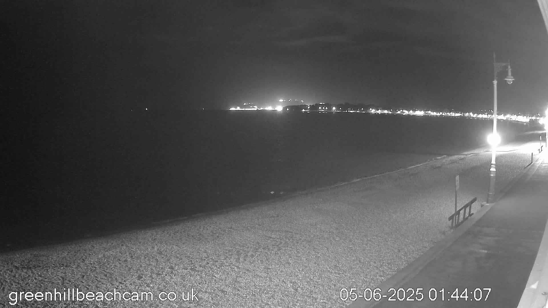 A dark, nighttime beach scene shows a pebbled shoreline with a lamppost and a distant town lit up across the water.