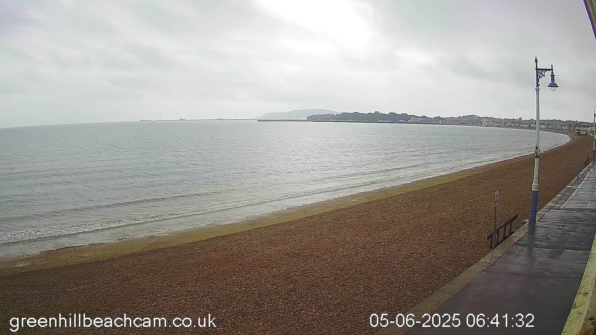 A pebble beach stretches along a calm, grey sea under an overcast sky, with a town visible in the distance.