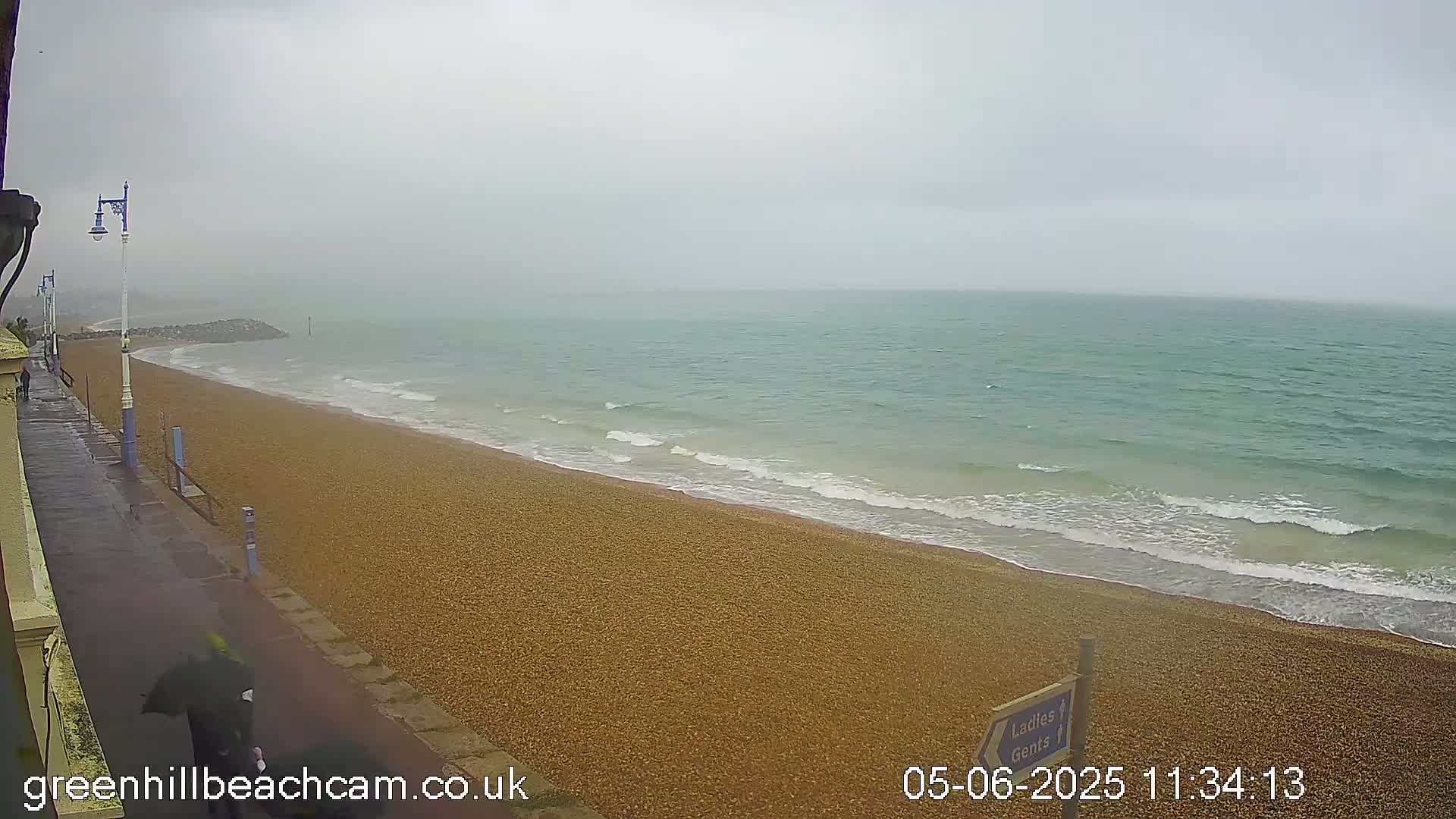 A person with an umbrella walks along a wet, pebbled beach next to a calm sea under an overcast sky.