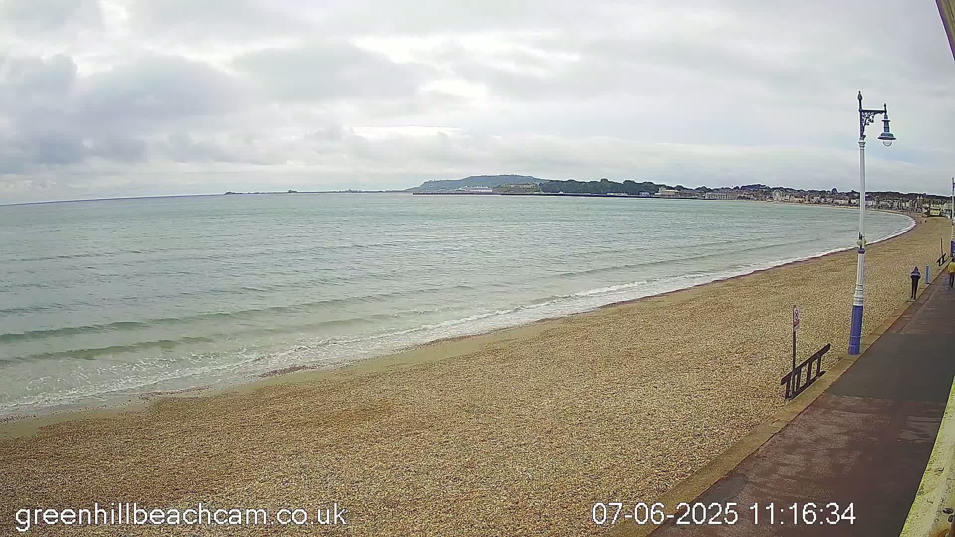 A sandy beach with gentle waves stretches along a calm sea under a cloudy sky, a promenade running alongside.