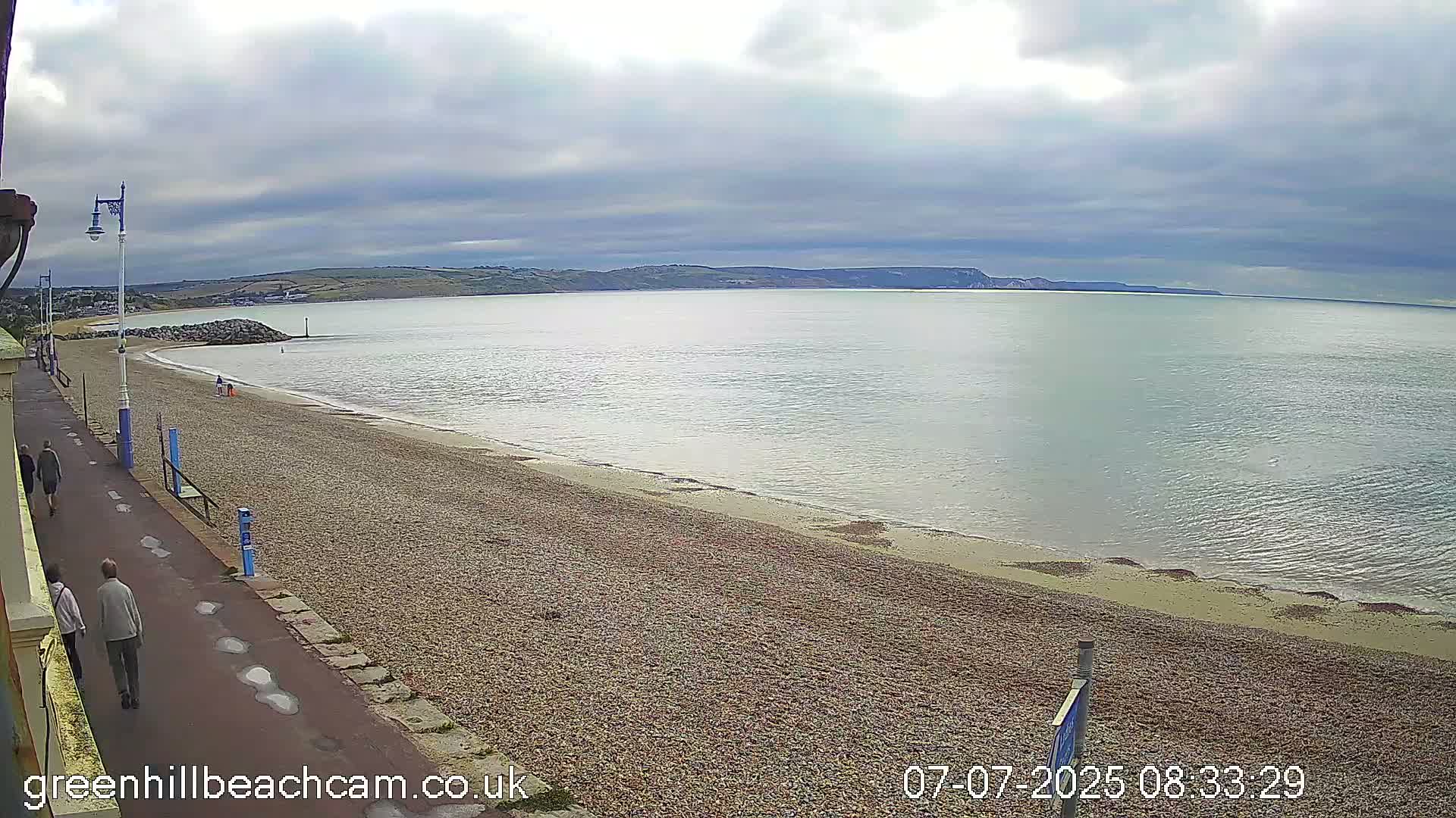 A pebble beach stretches alongside a calm sea under a cloudy sky, with a few people walking along a paved path.