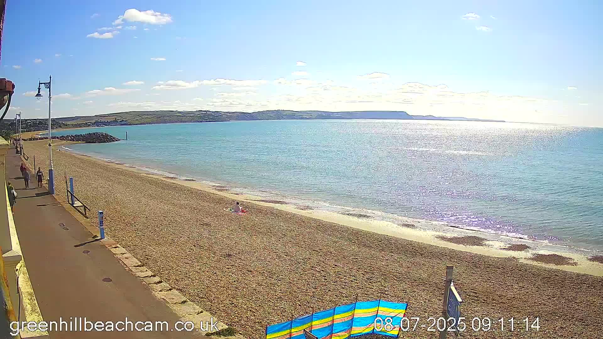 A pebble beach stretches along a calm, sunny sea under a mostly clear blue sky, with a few people walking on the promenade and some relaxing on the sand.