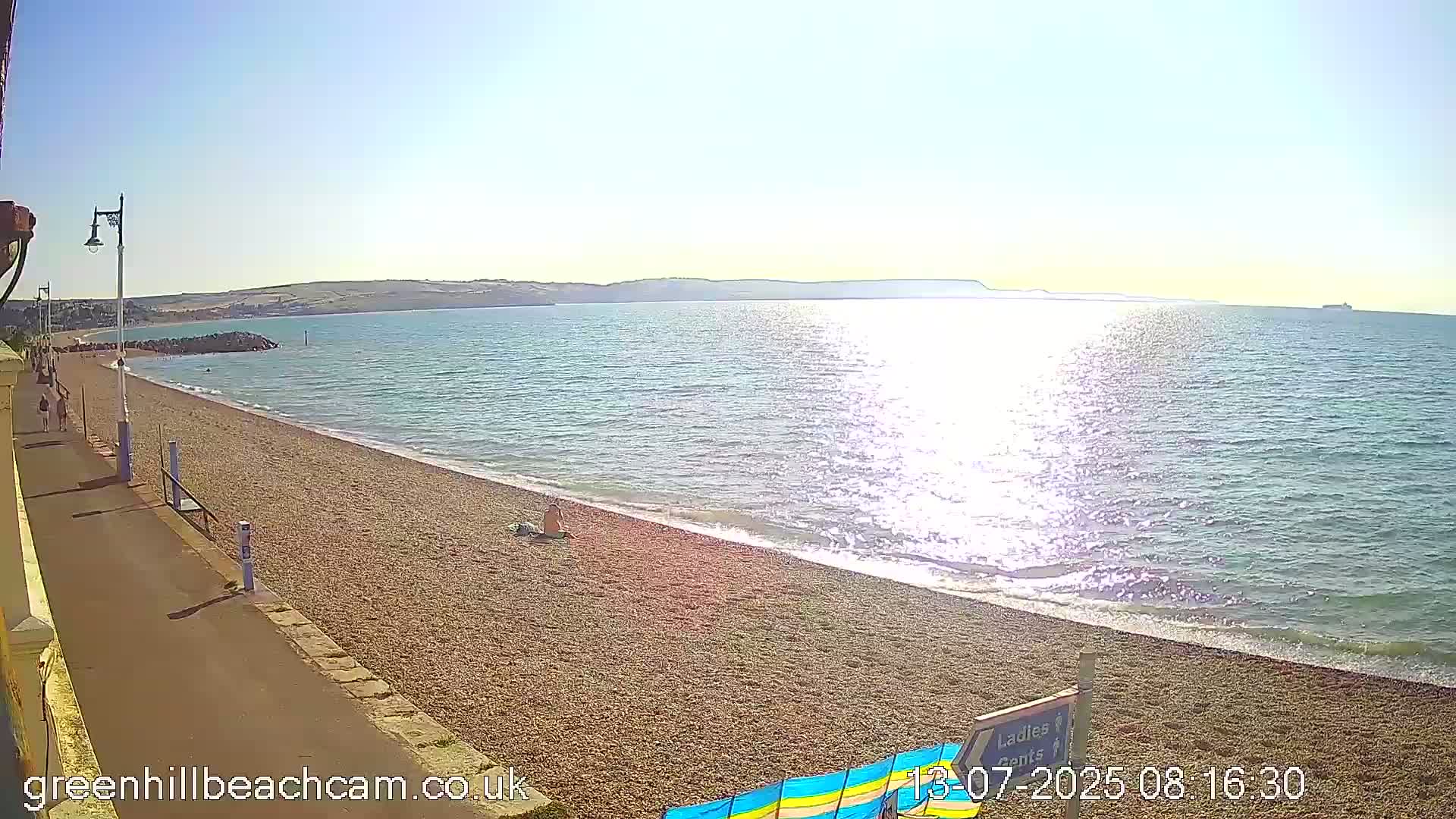 A sunny day at a pebble beach shows calm water reflecting sunlight, with a paved walkway and distant hills in view.