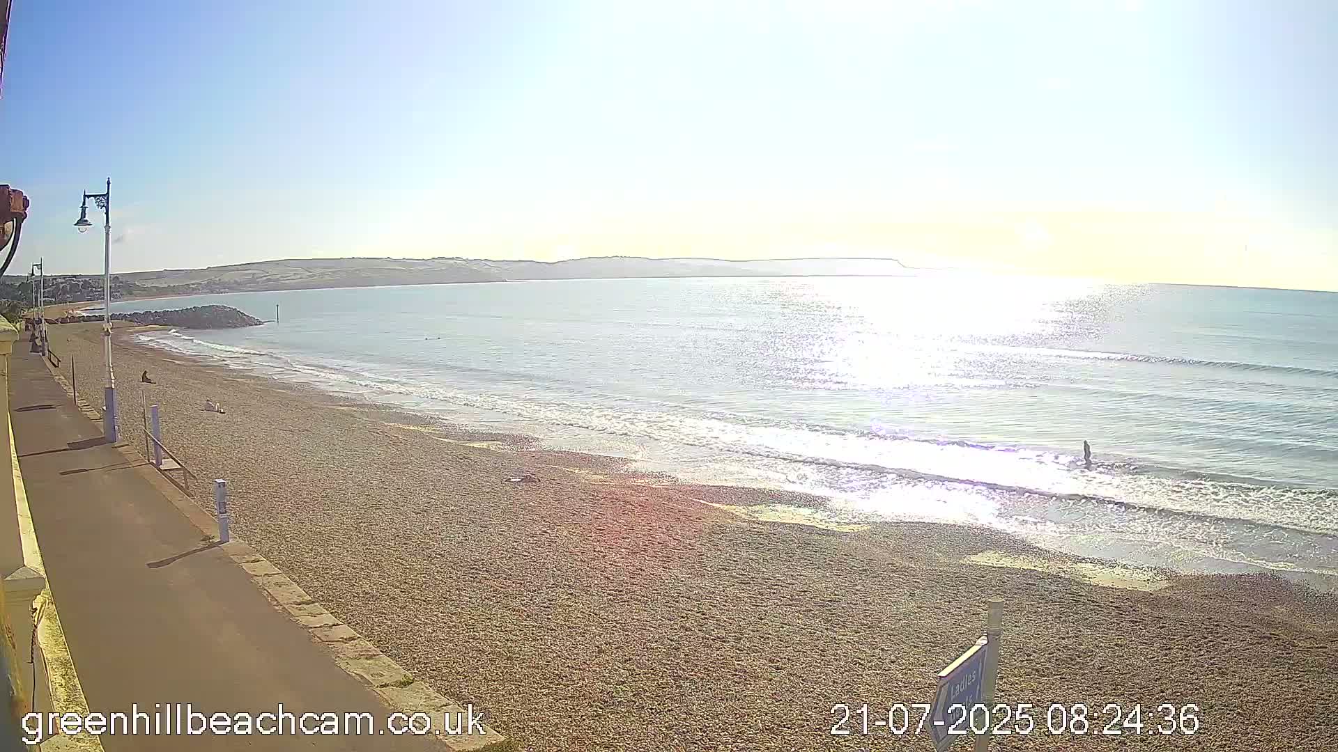 A sunny day at a pebble beach shows calm waves lapping the shore, a paved walkway, and a few people on the beach.
