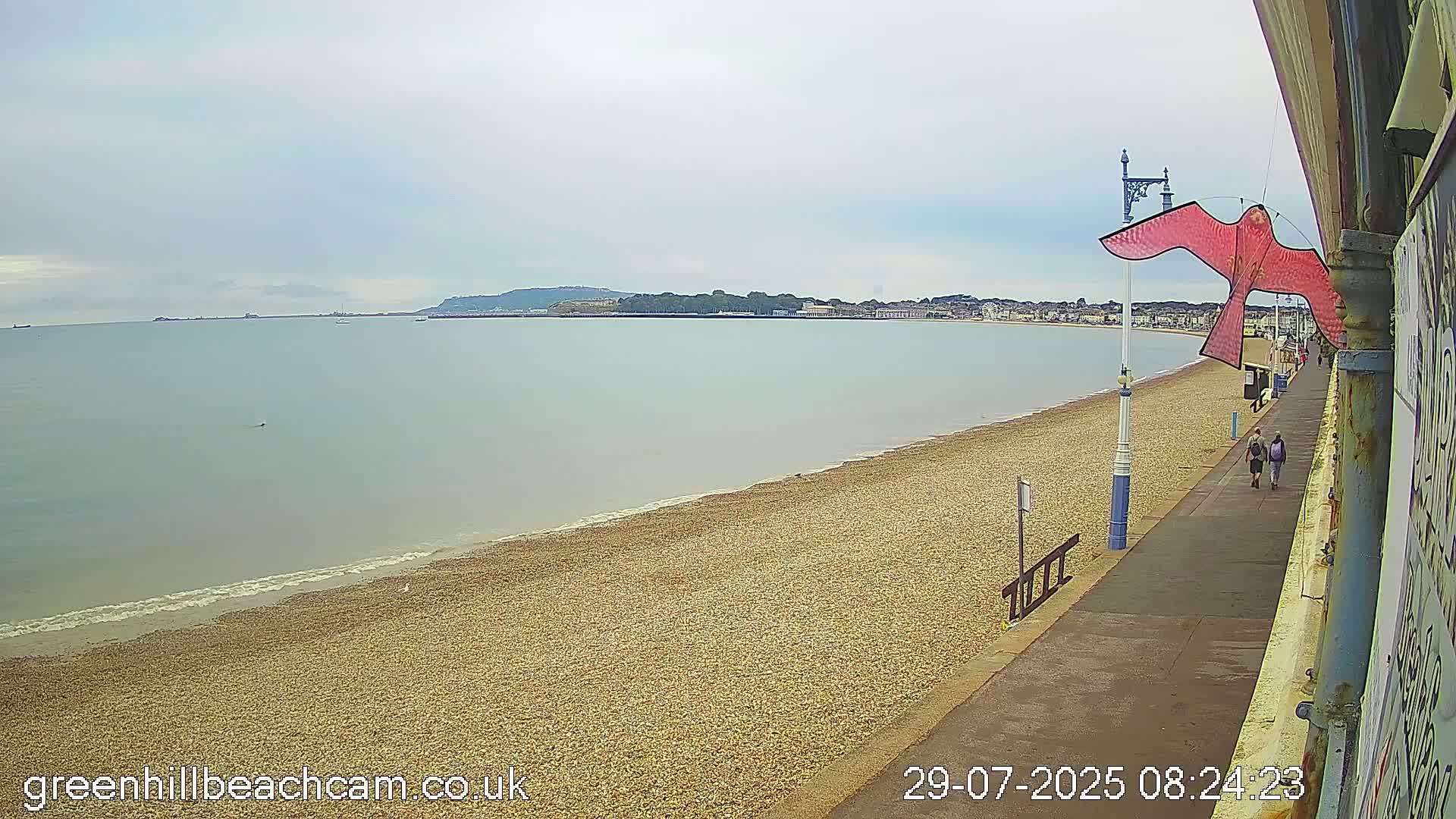 A calm, overcast day reveals a pebbled beach stretching to a calm sea, with a coastal town and hills visible in the distance, and two people walking along a promenade.