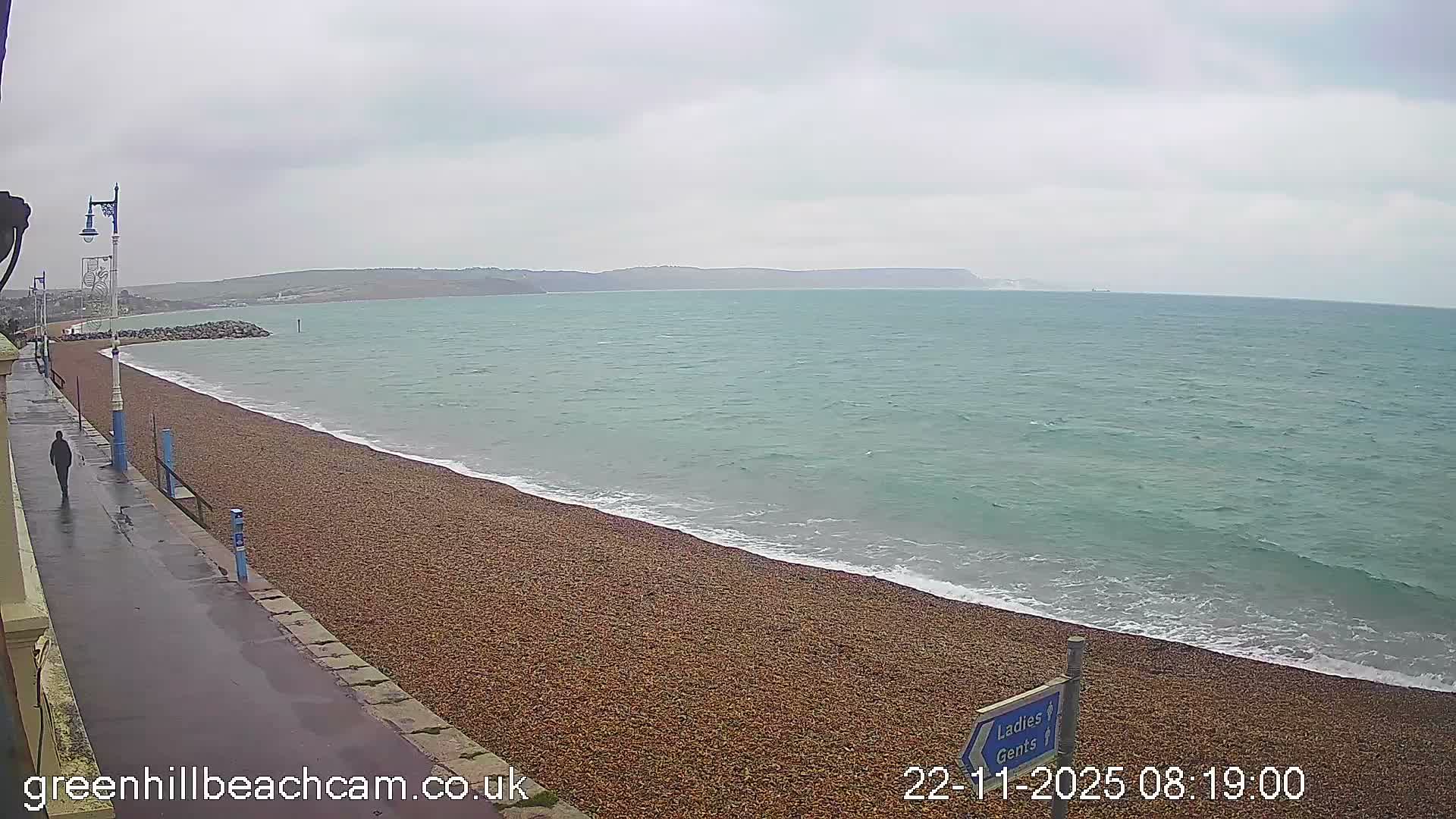 A solitary person walks on a wet promenade bordering a pebble beach and the turquoise ocean, with a rocky breakwater and distant hills under an overcast sky.