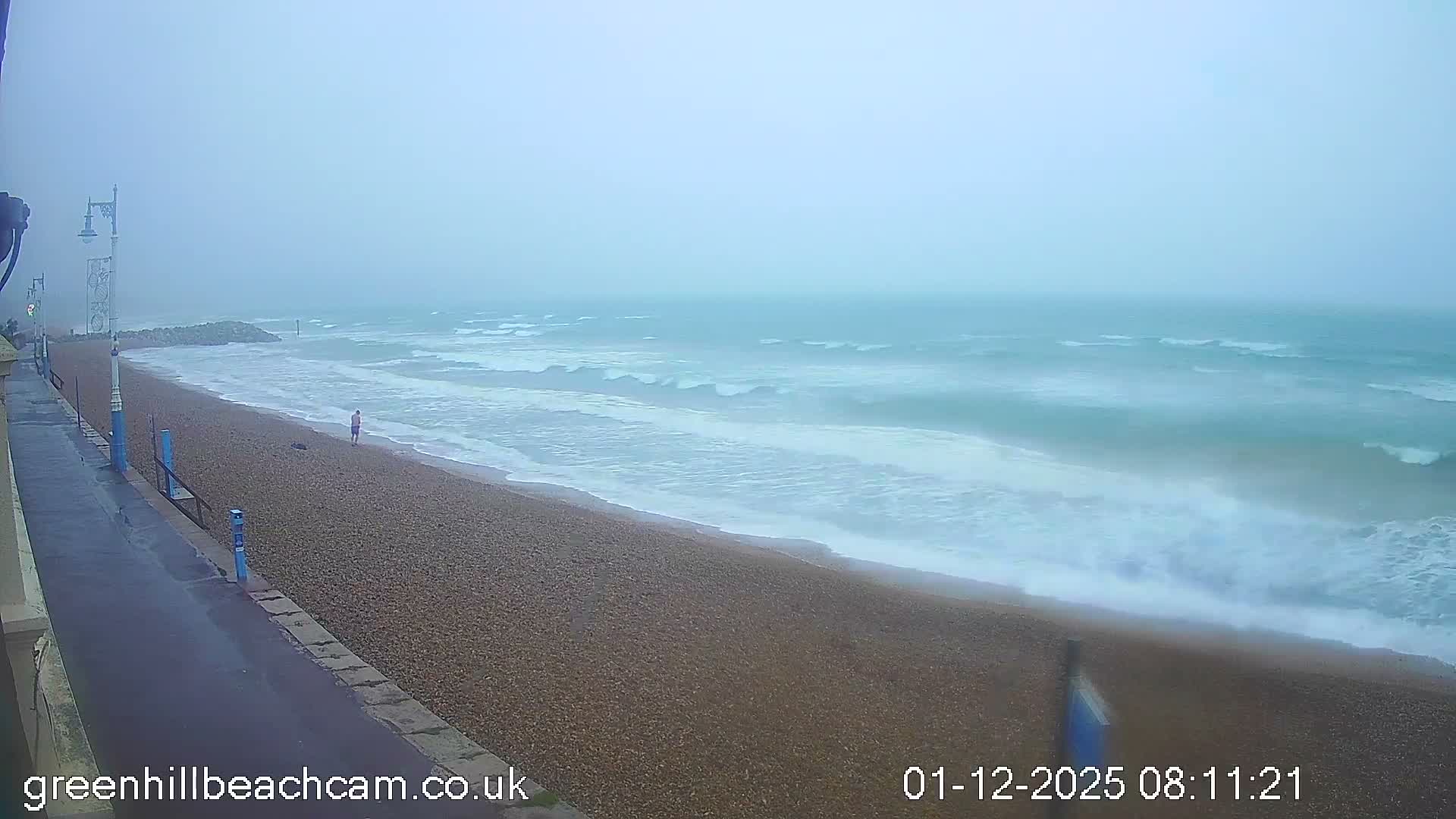 A misty, overcast day overlooks a pebbled beach where a lone person walks near active waves breaking on the shore, with a concrete promenade running along the left side.