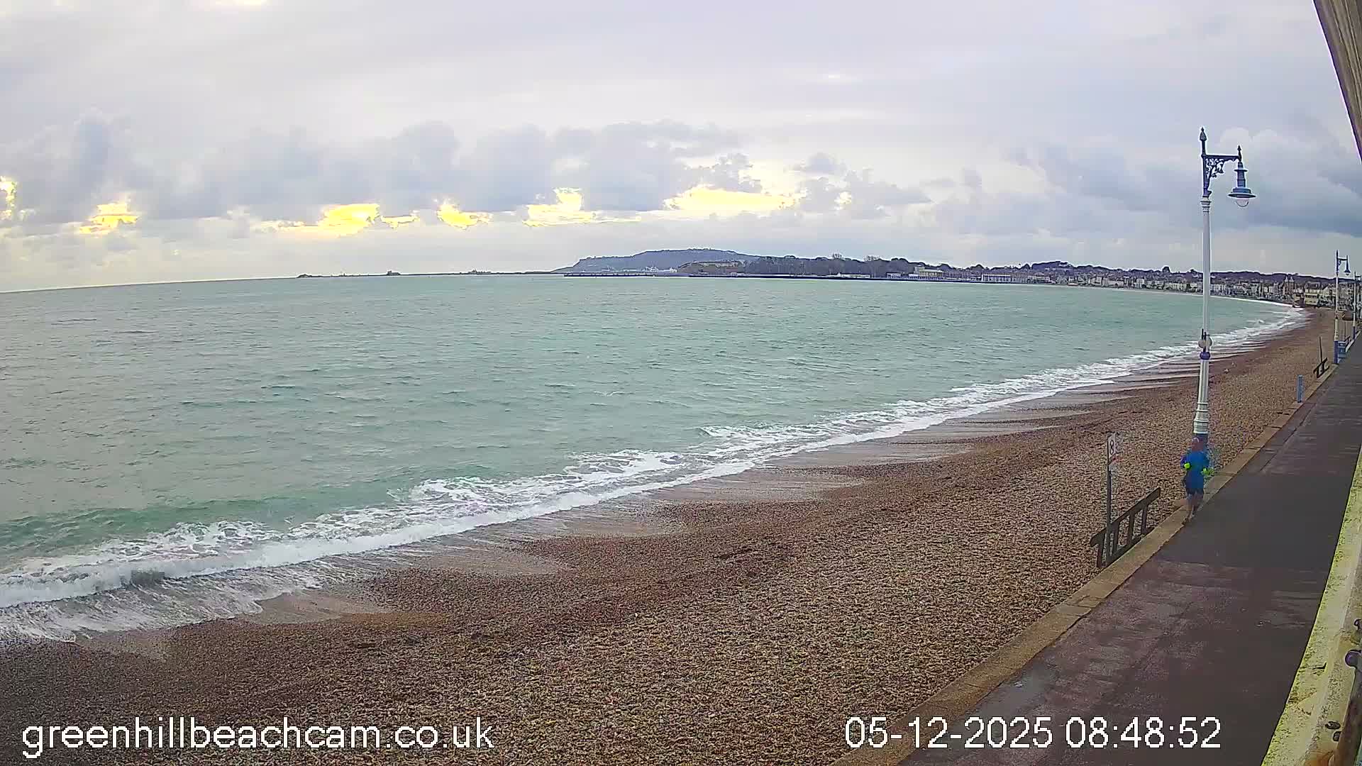 A wide view captures a pebble beach with gentle waves under a cloudy morning sky with hints of yellow light, a person jogging on the adjacent promenade, and a distant town and headland along the curving coastline.