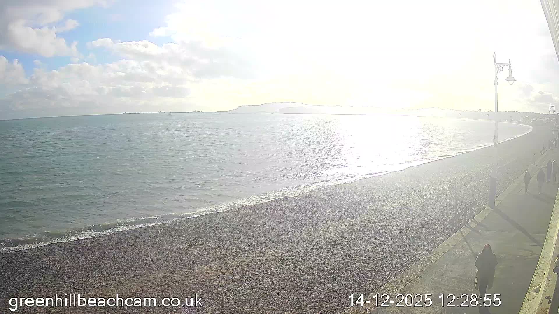 A wide view captures a pebble beach with gentle waves under a cloudy morning sky with hints of yellow light, a person jogging on the adjacent promenade, and a distant town and headland along the curving coastline.