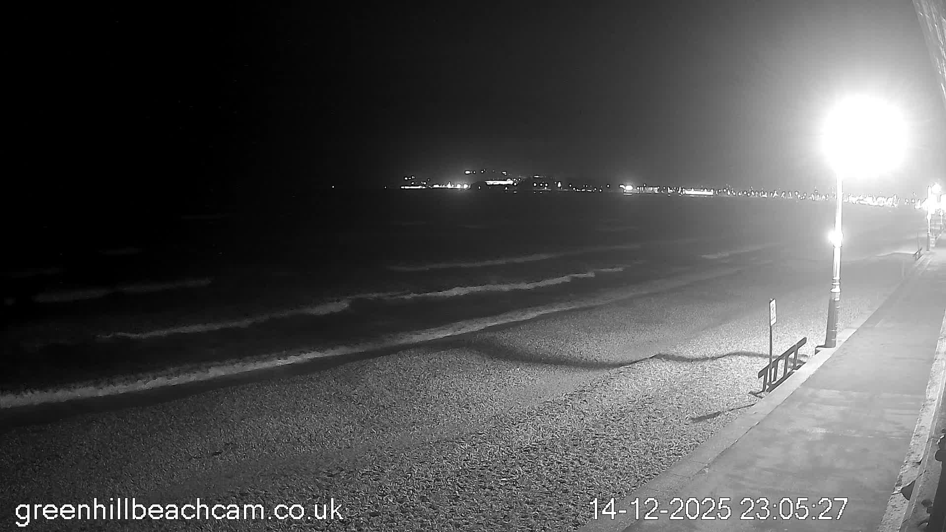 A wide view captures a pebble beach with gentle waves under a cloudy morning sky with hints of yellow light, a person jogging on the adjacent promenade, and a distant town and headland along the curving coastline.